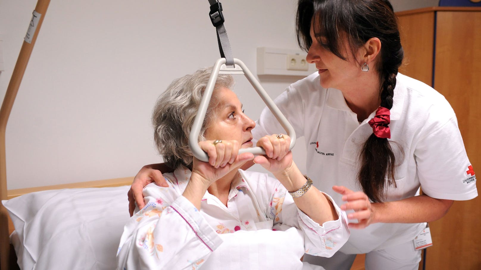 Eine Patientin betreut von einer Diplomkrankenschwester hält sich an einer Triangel-Aufrichtungshilfe fest. Symbolbild