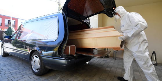 Ein Bestatter in Schutzkleidung schiebt einen Sarg in einen Leichenwagen. Symbolbild