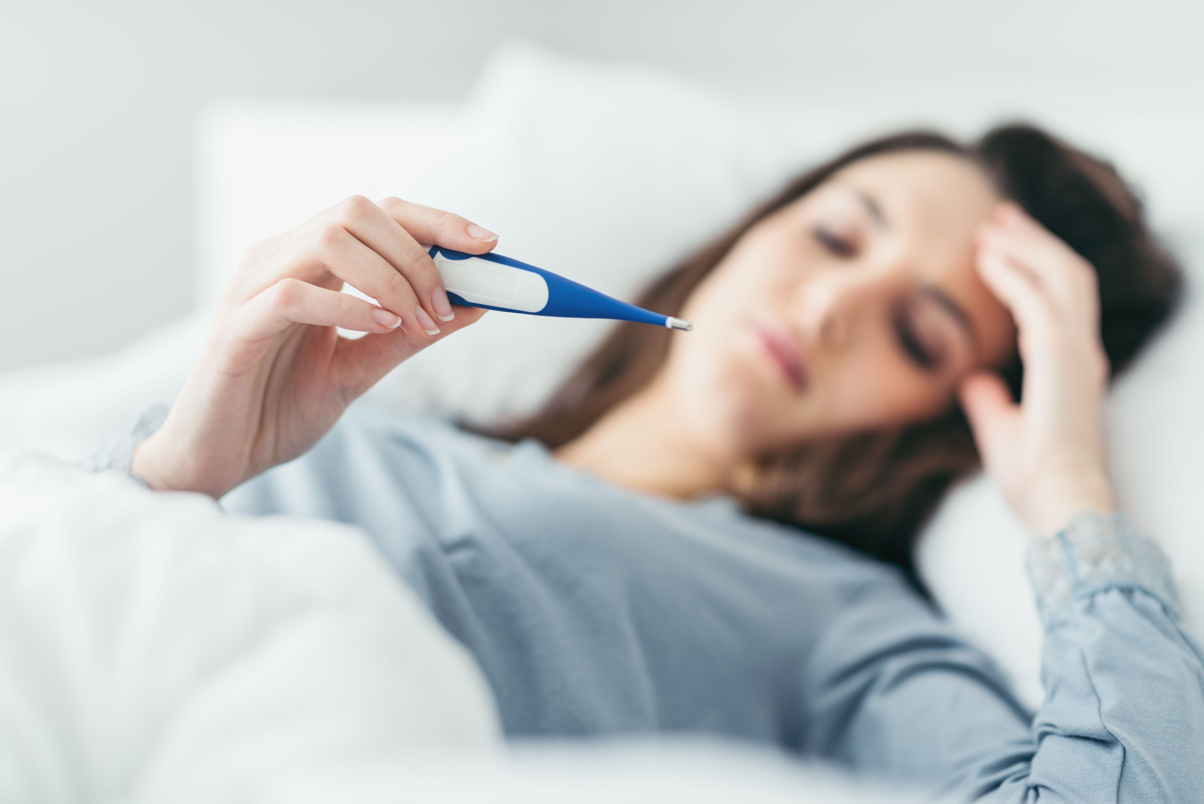 Woman with flu virus lying in bed, she is measuring her temperature with a thermometer and touching her forehead