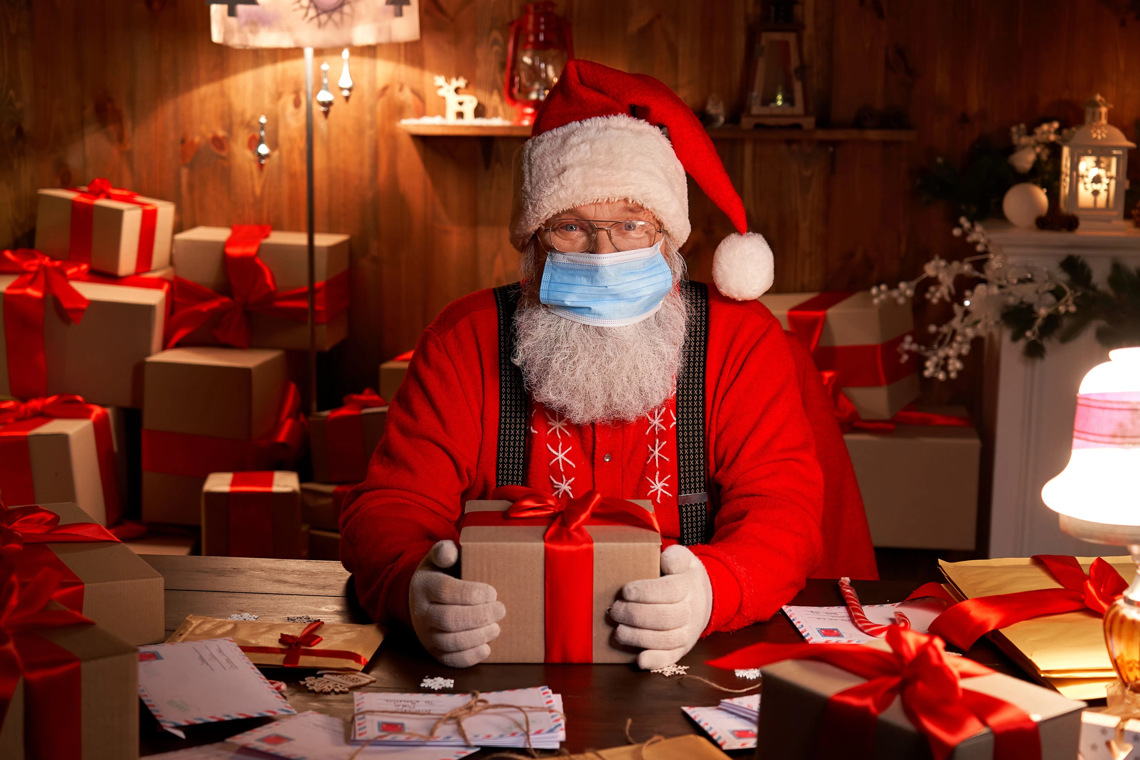 Old funny bearded Santa Claus wearing face mask, holding gift box preparing for xmas eve sitting at cozy home table late in night with presents. Merry Christmas Covid 19 coronavirus safe delivery.