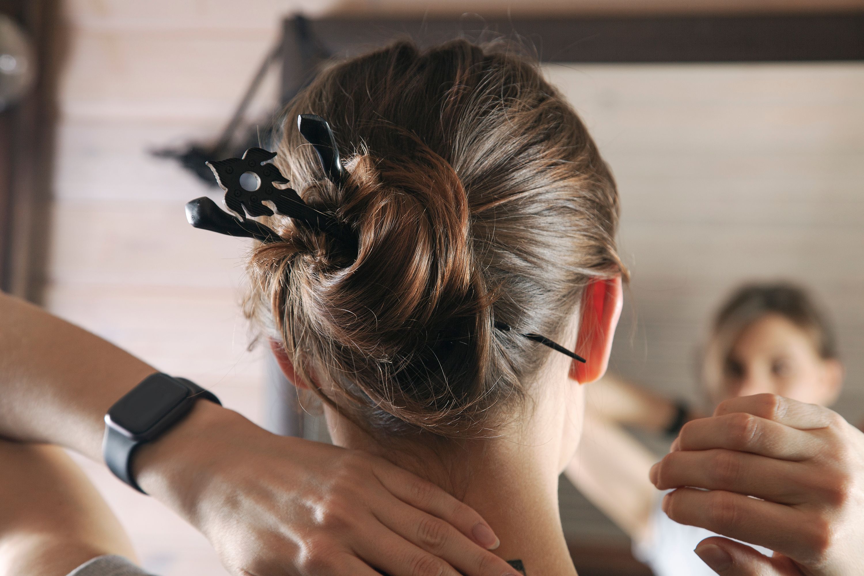 Back view of young attractive woman with hair bun shell-wrapped, with three wooden hairpins of sophisticated shapes. Close up head, hand with black electronic watch on wrist, reflection in the mirror.