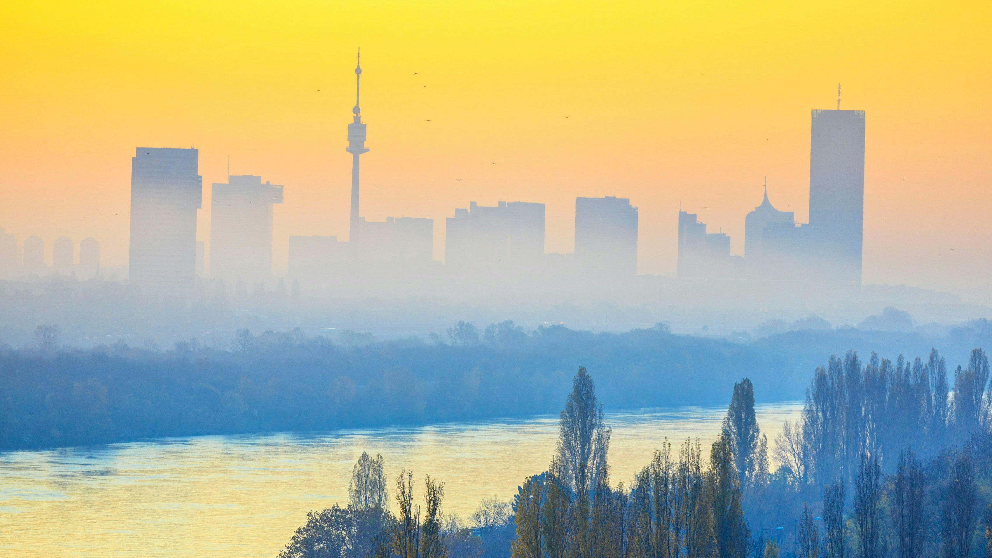 Blick kurz nach Sonnenaufgang vom Leopoldsberg hinunter über die Donau und auf die aus dem Dunst ragenden Hochhäusern der Stadt Wien.