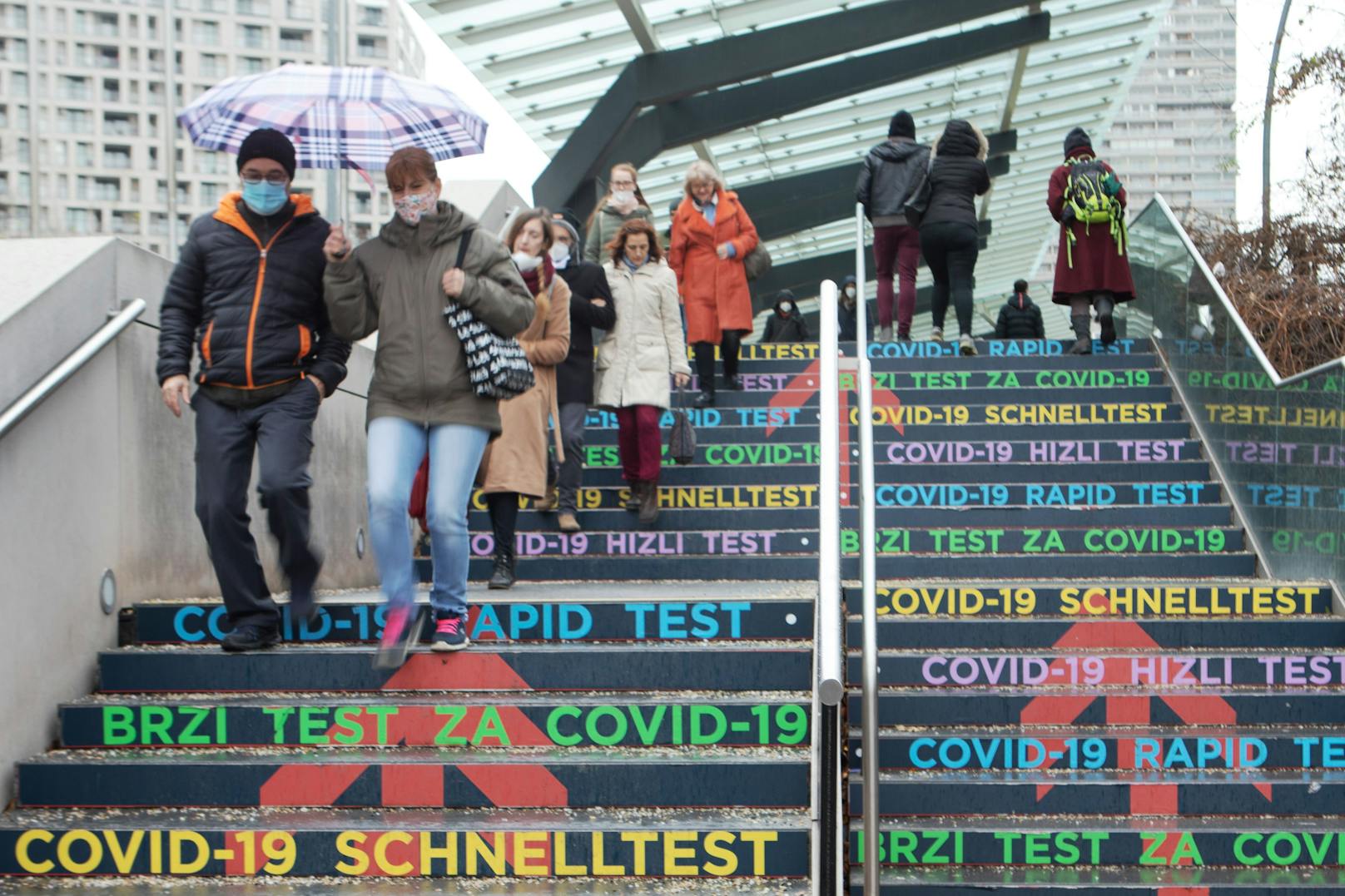 Menschen tragen Schutzmasken und Regenschirm am Weg zu dem Antigen Schnelltest an einer Stiege bei dem Austria Center Vienna in Wien.
