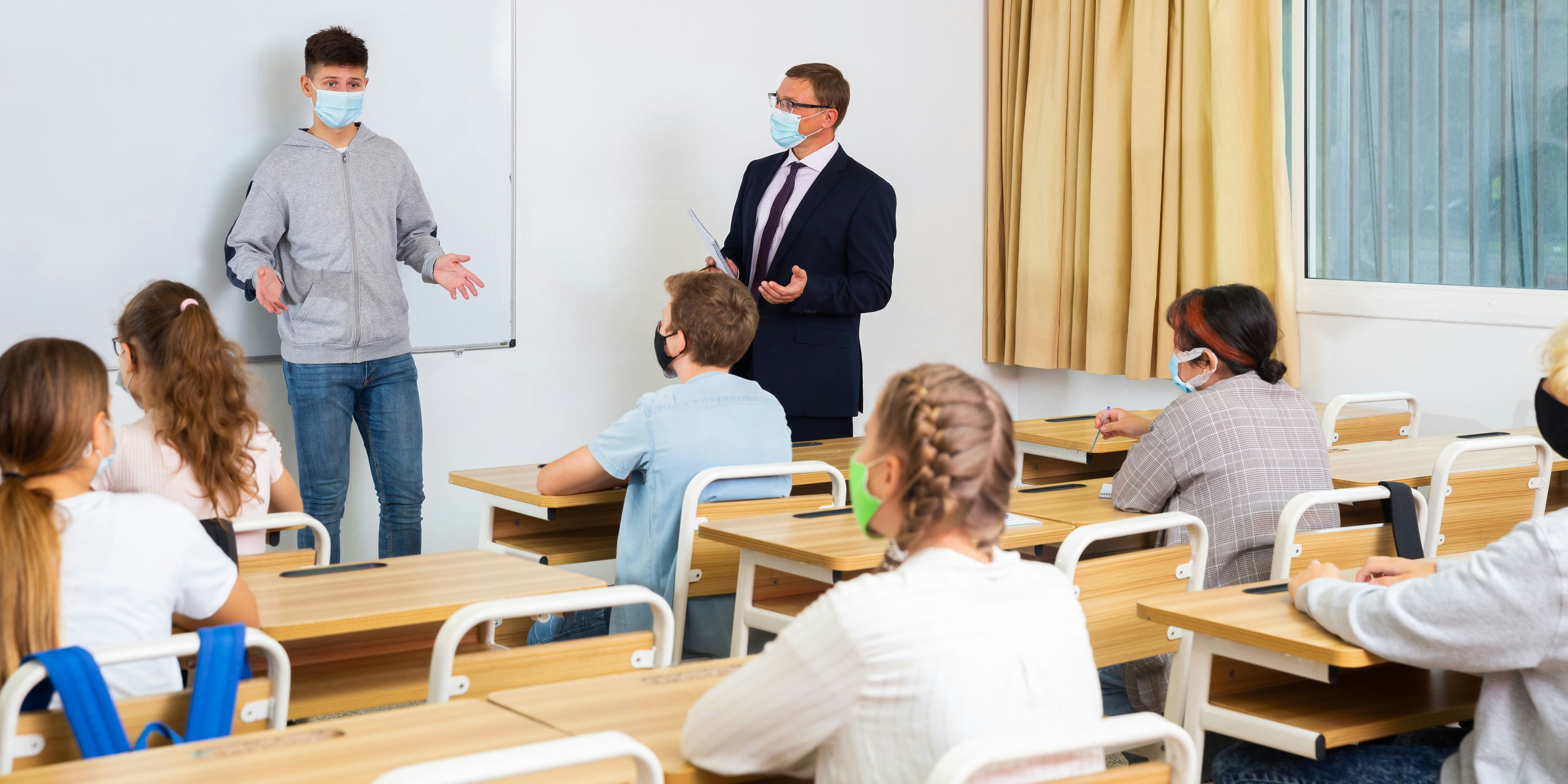 Portrait of diligent teenager schoolboy wearing protective face mask answering at board to teachers question in front of group of students in classroom, new normal education during pandemic