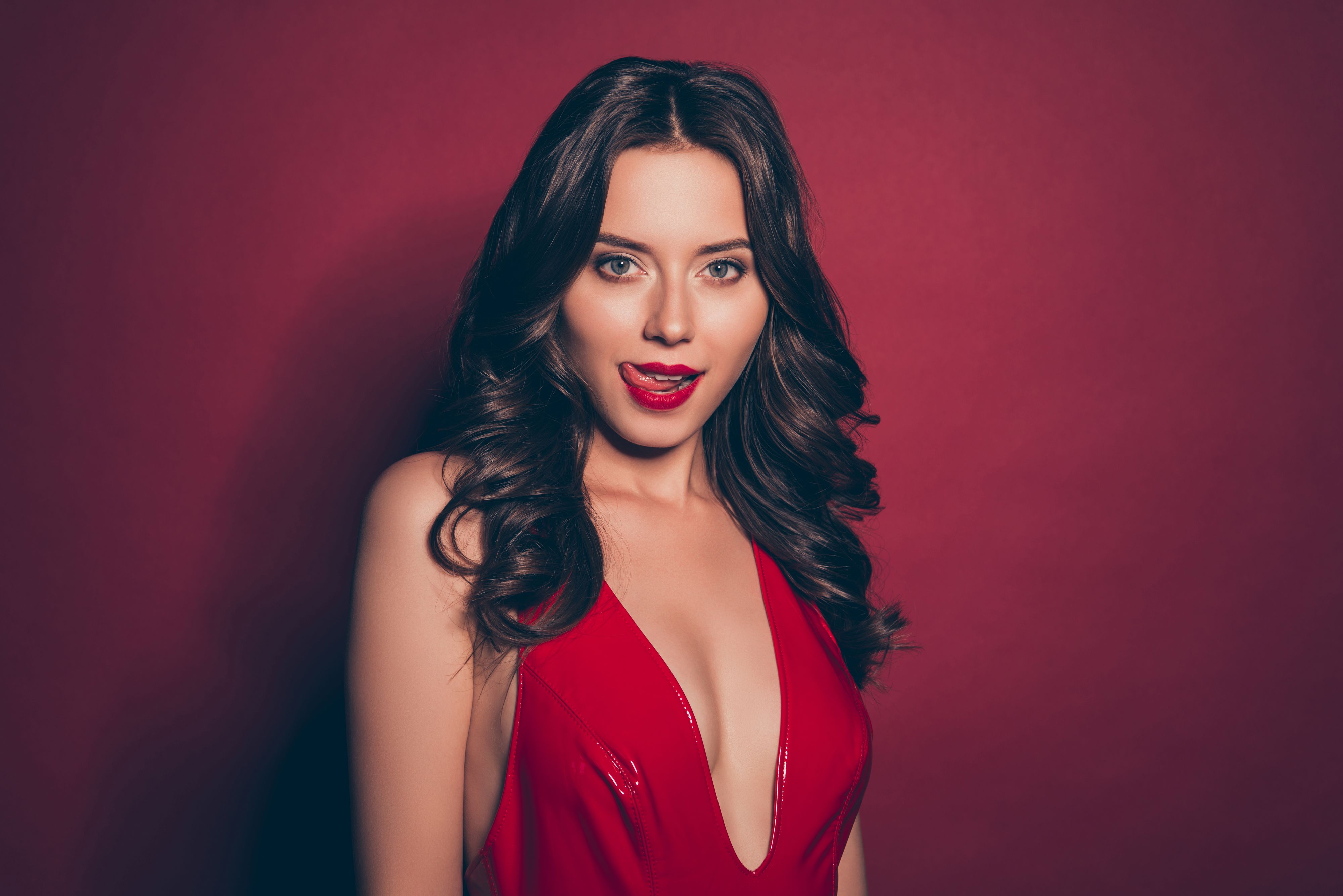 Good-wearing alluring pretty brunette lady with modern curly wavy hairdo look at camera show tongue out lick lips she stand in her lustrous classy shine mini wear isolated on deep red background