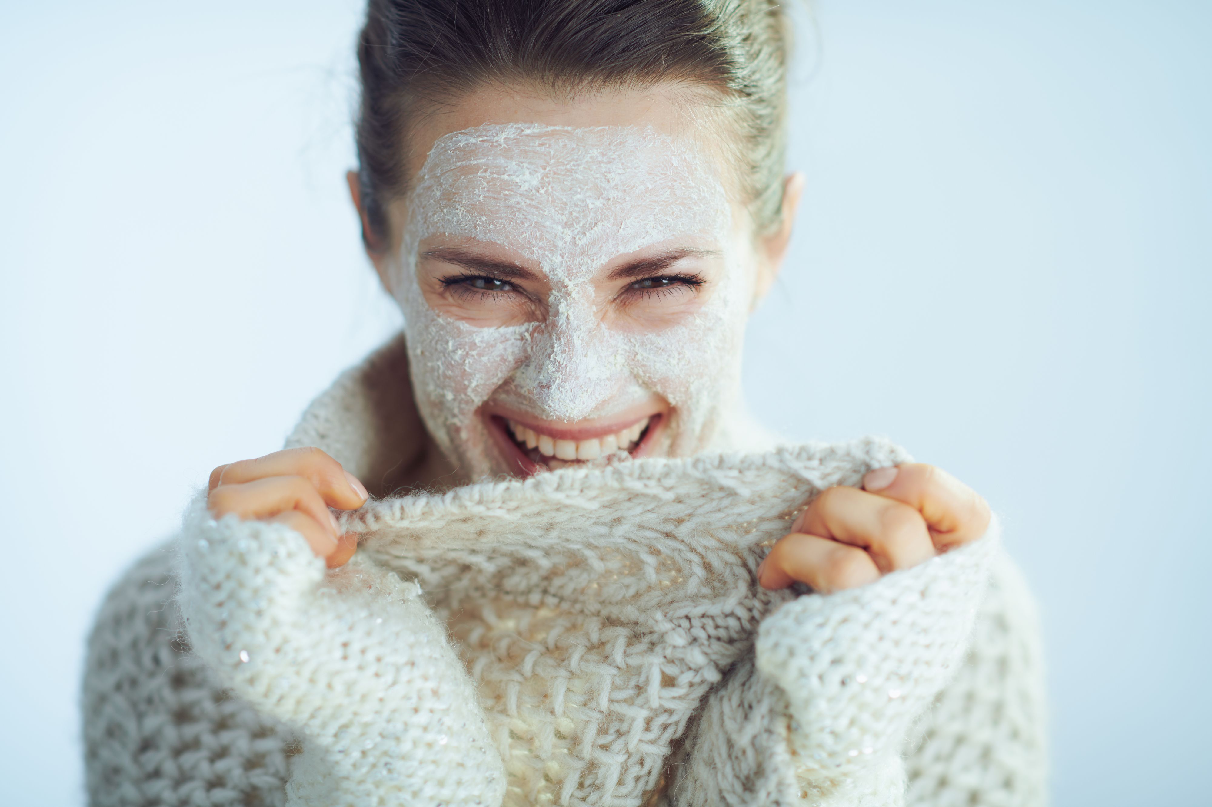 happy modern woman in roll neck sweater and cardigan with white facial mask as part of winter skin care playing with clothes on winter light blue background.