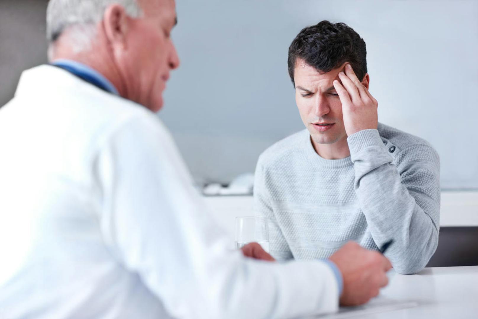 Young man rubbing his forehead in agony while his doctor is writing out a prescription