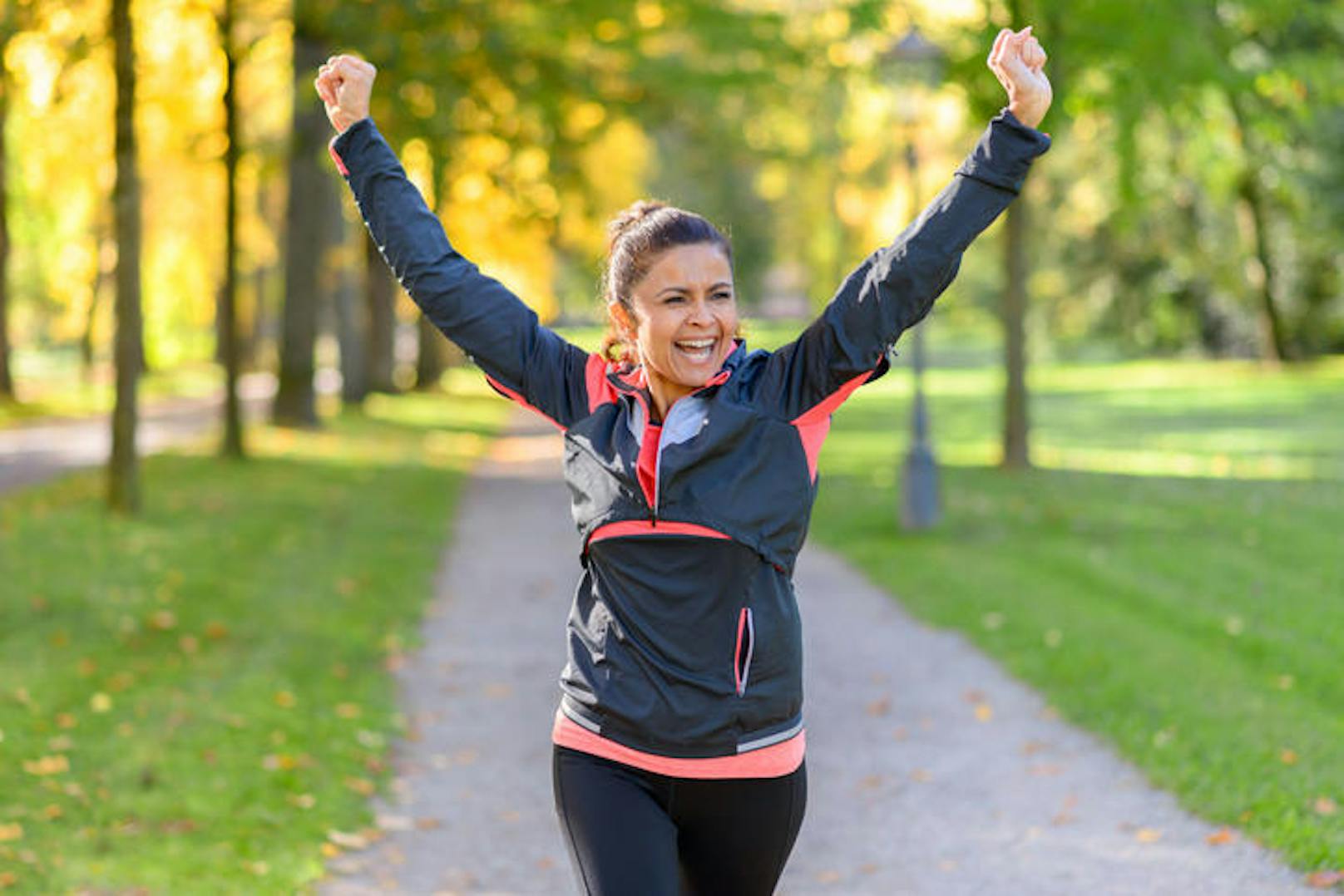 Happy fit middle aged woman cheering and celebrating as she walks along a rural lane through a leafy green park after working out jogging (Happy fit middle aged woman cheering and celebrating as she walks along a rural lane through a leafy green park 