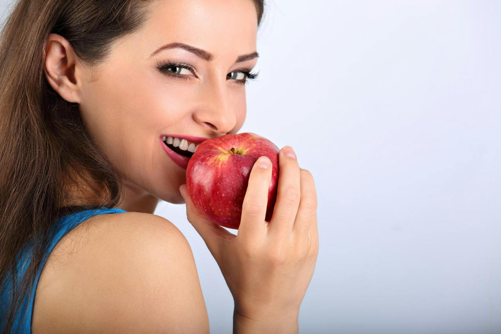 Beautiful excited makeup brunette woman biting the red tasty apple and looking happy with empty space blue background. Closeup portrait (Beautiful excited makeup brunette woman biting the red tasty apple and looking happy with empty space blue backgro