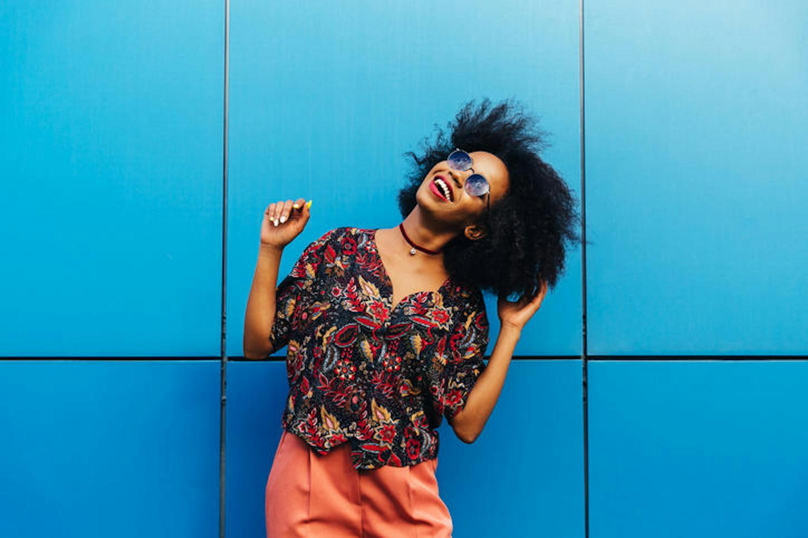 Cheerful attractive afro american girl enjoying spending time outdoors, dancing, wearing sunglasses, dressed in colorful blouse and pants, looking at the sky. On blue wall background (Cheerful attractive afro american girl enjoying spending time outdo