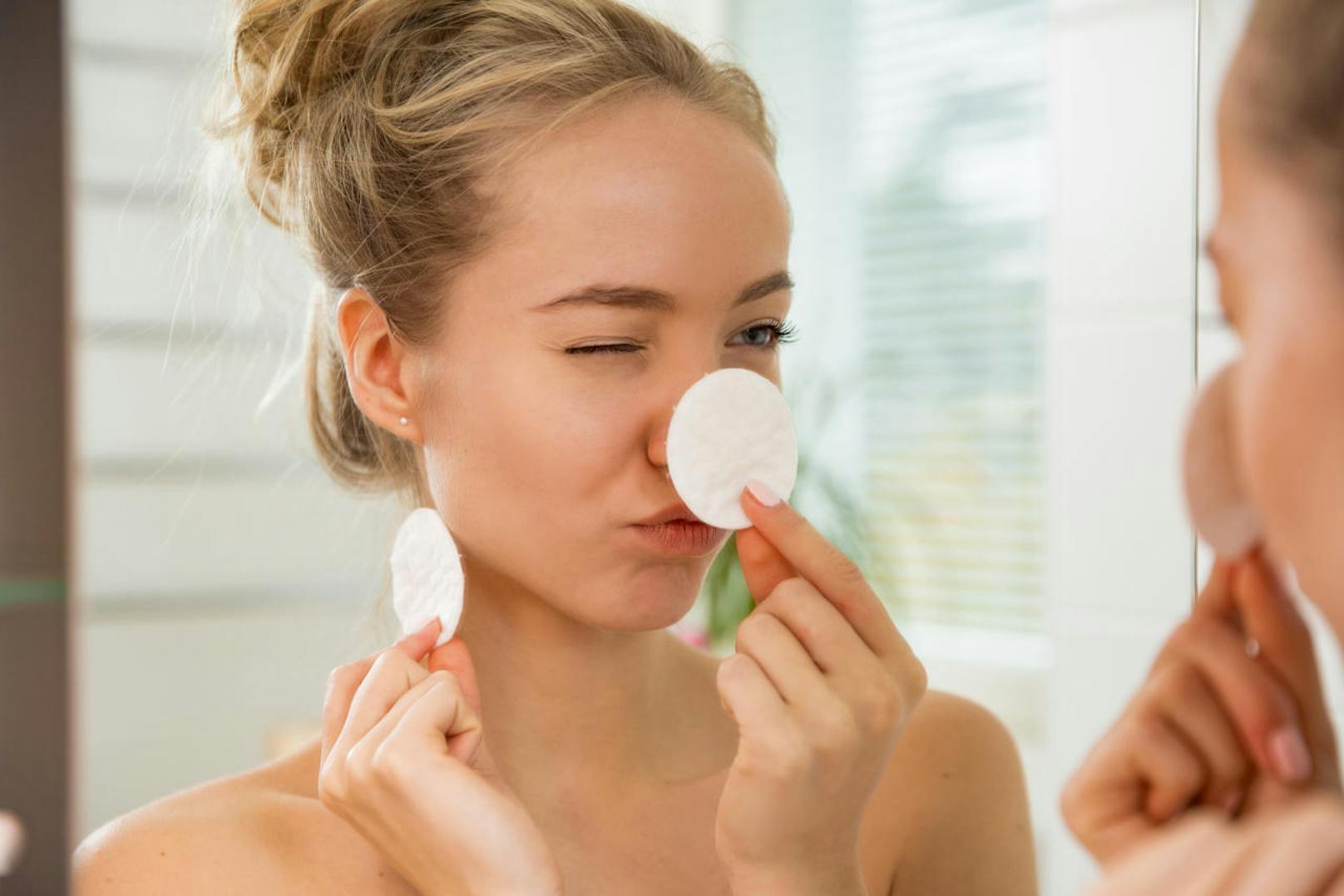 Young beautiful woman cleaning her face skin with cotton pad in bathroom. Standing in towel, looking in the mirror, laughing and having fun. Morning skincare routine.
