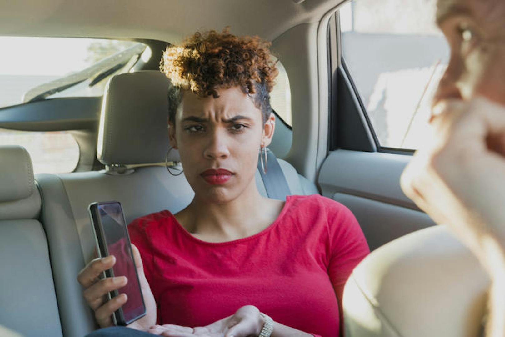 This is a horizontal, color photograph of a young mixed race, black Millenial woman riding as a passenger in the backseat of a rideshare taxi in Miami, Florida. She is angry and shows the driver her phone. He looks back upset and worried with his bad directions. Photographed with a Nikon D800 DSLR camera in natural light.