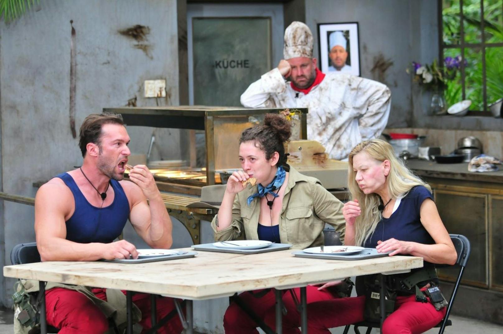 Bastian Yotta, Leila Lowfire und Sibylle Rauch holen sich an der Theke ihr Stück von der Dschungelpizza, nehmen am Tisch Platz und die Zeit läuft.