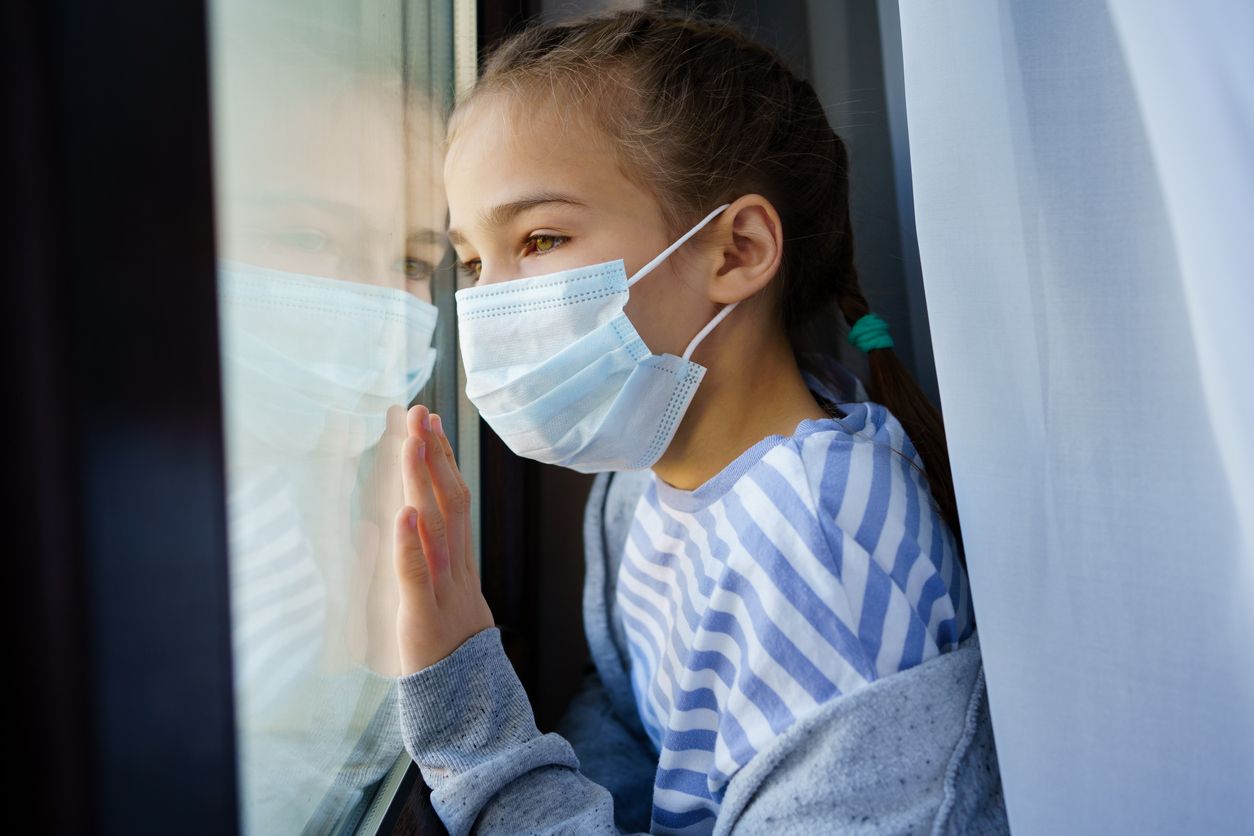 a teen girl in a protective mask, look out of the window outside. she is sick and self-isolating during the pandemic. misses her friends and parents. distance learning.