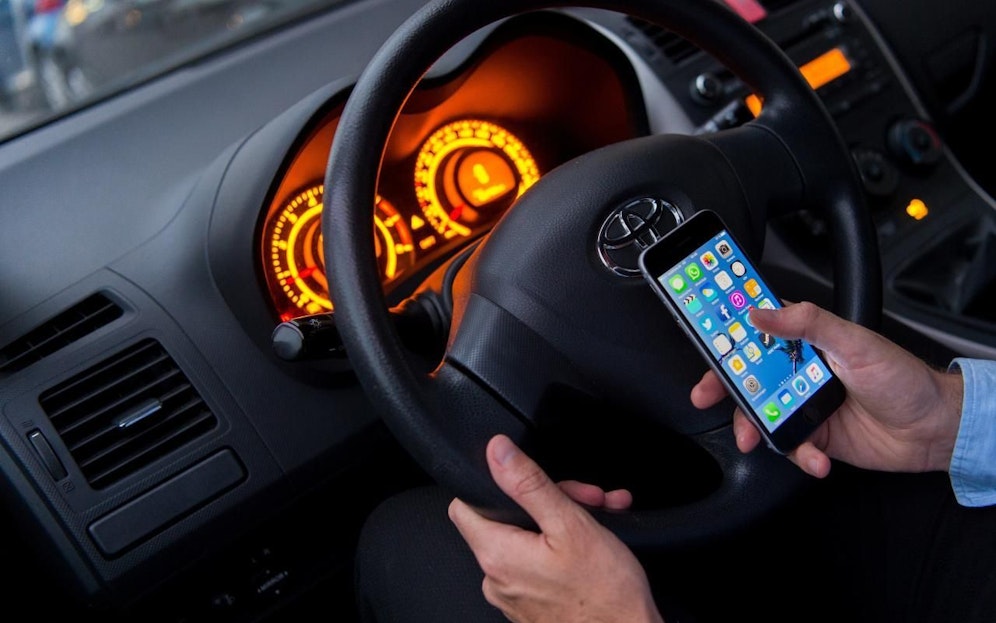 Ein Mann sitzt in einem Auto am Steuer und hält ein Smartphone in der Hand. Symbolfoto