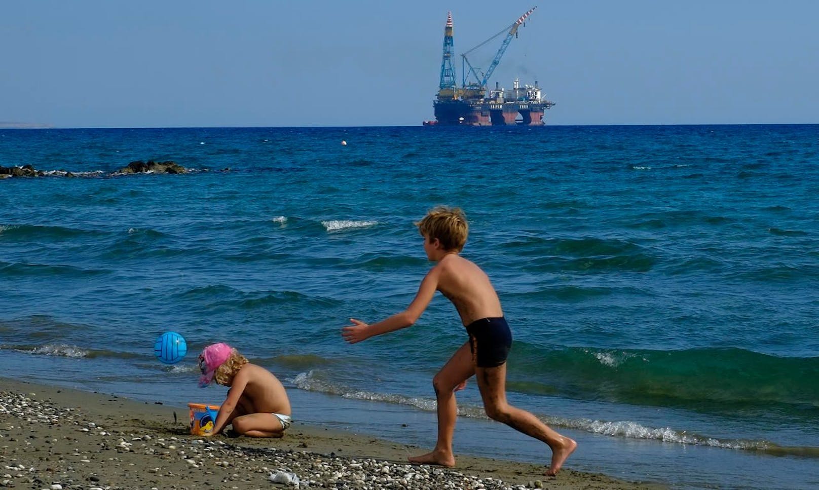 Kinder spielen an einem Strand am Stadtrand von Larnaca. Im Hintergrund steht eine Bohrplattform.