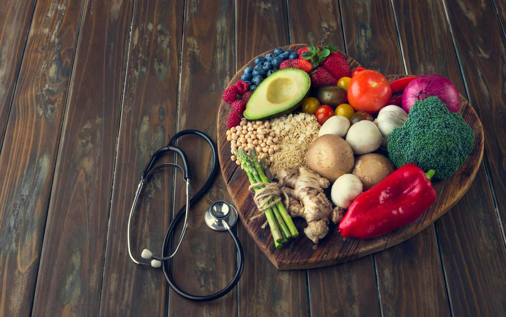 Healthy food on a heart shape cutting board. Love of food concept with fruit, vegetables, grains and high fibre foods. Rustic wood textures. There is also a stethoscope 