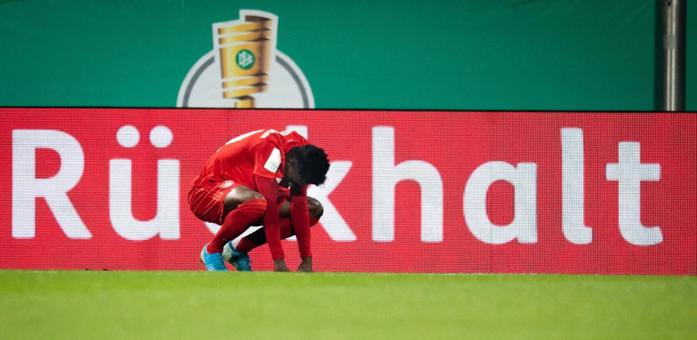 Alphonso Davies hockelt nach seinem Eigentor niedergeschlagen am Feld. Bayern-Fans regt auf, dass ihn keiner tröstet. Die Bandenaufschrift "Rückhalt" sorgt für eine Portion Ironie.