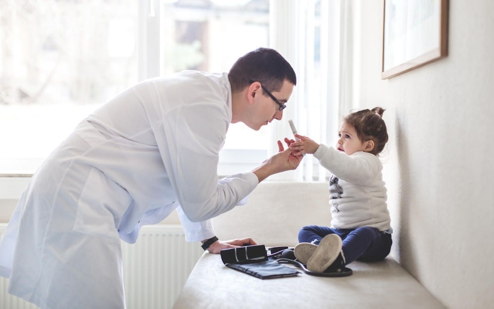 Doctor and toddler girl are inside patient's room. The girl is holding medical light and is checking up on doctors eye site.