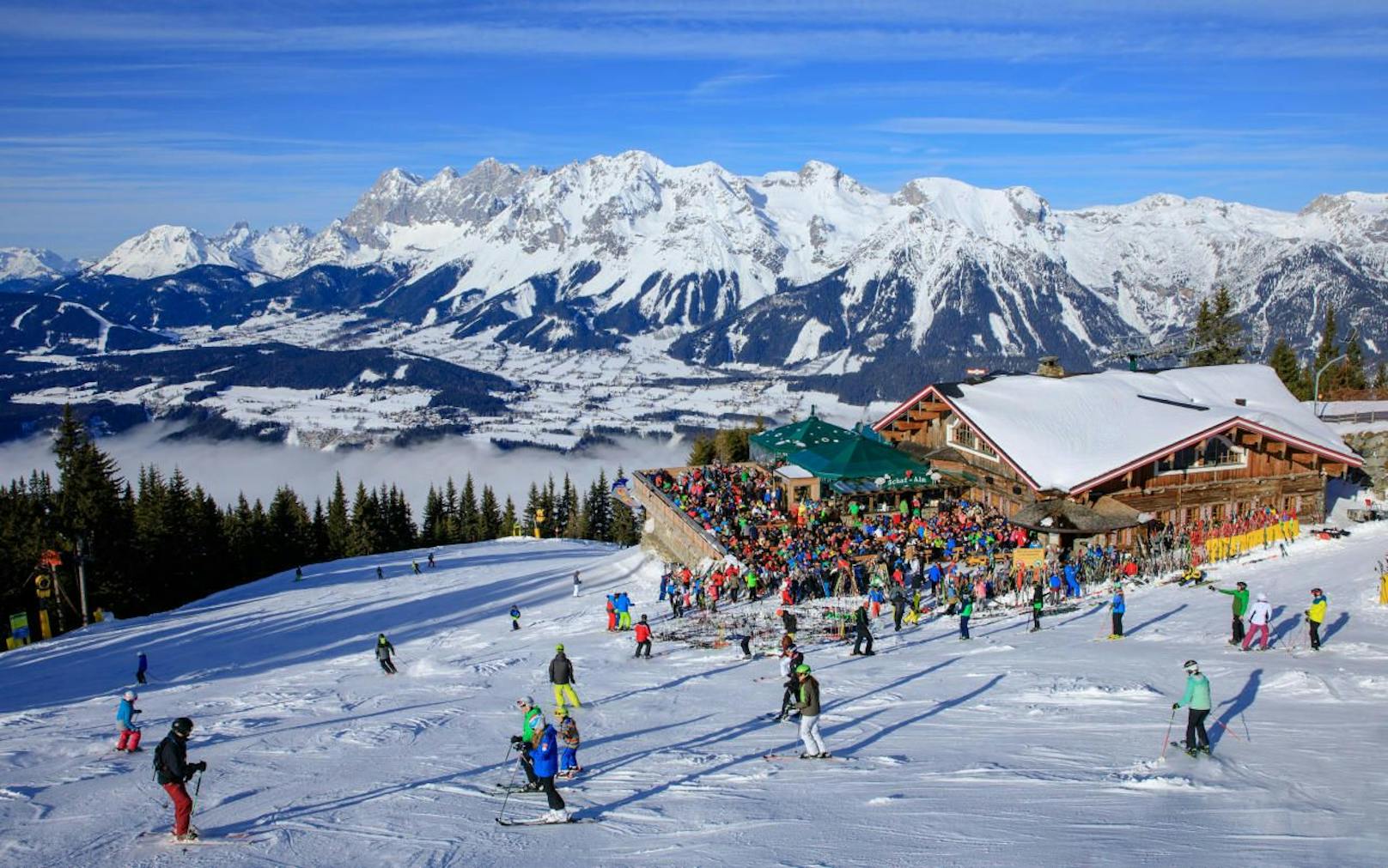 Skifahrer auf der Piste und auf der Terrasse der Skihütte Schafalm vor der Kulisse des Dachstein-Gebirges. Symbolbild