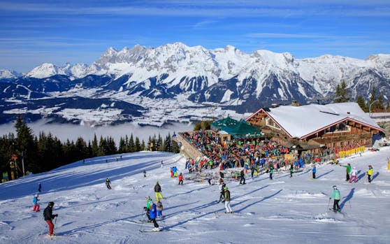 Skifahrer auf der Piste und auf der Terrasse der Skihütte Schafalm vor der Kulisse des Dachstein-Gebirges. Symbolbild