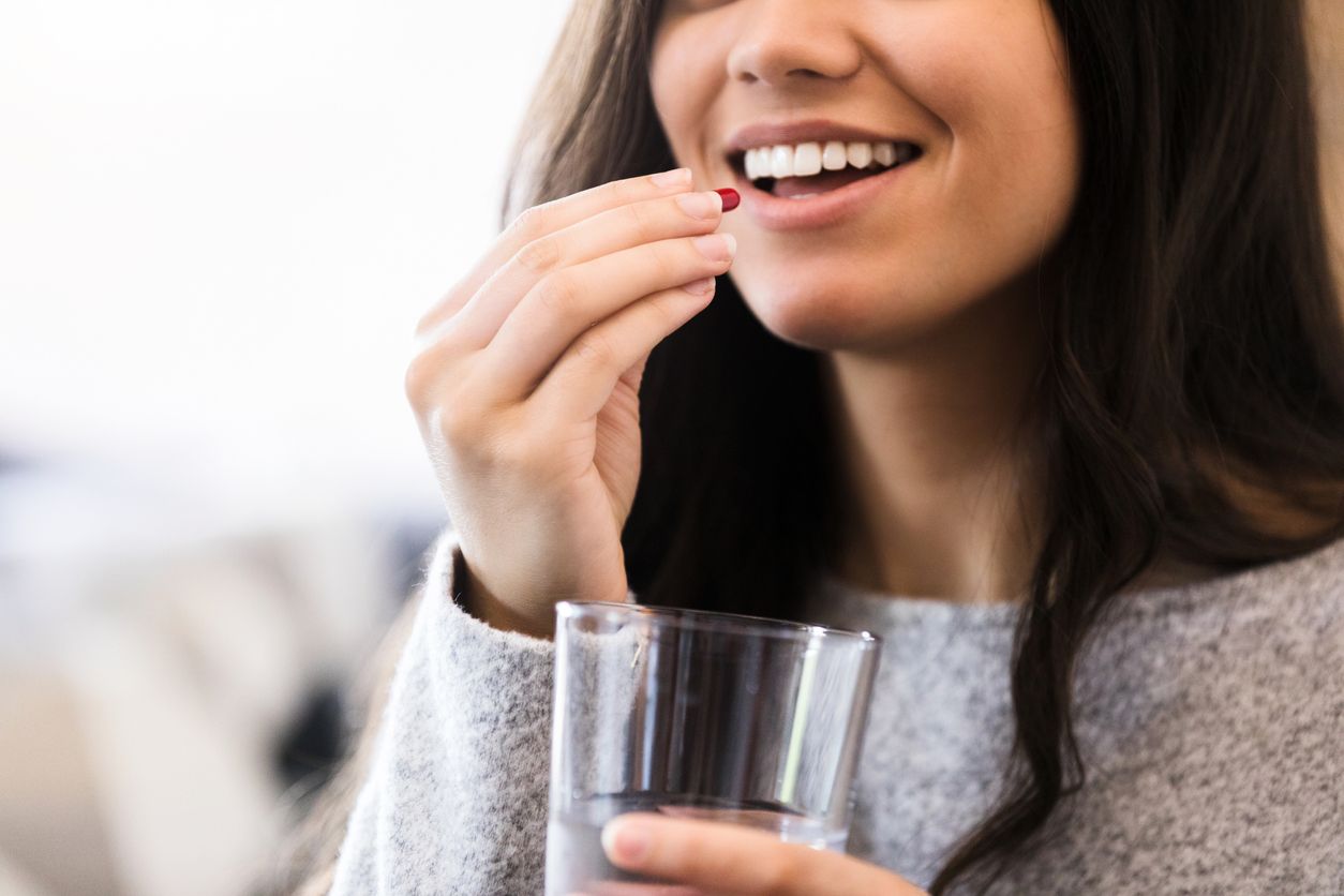 A confident young woman smiles as she takes a daily nutritional supplement. She is holding a water glass.