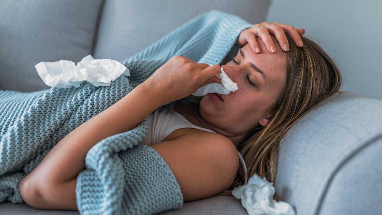 Sick Woman having Flu with Caught Cold. Sneezing into Tissue. Headache. Virus. Sick woman lying in bed with high fever. She is blowing nose. Photo of Young female Lying On Bed covered with a blanket  Infected With Allergy holding her head.