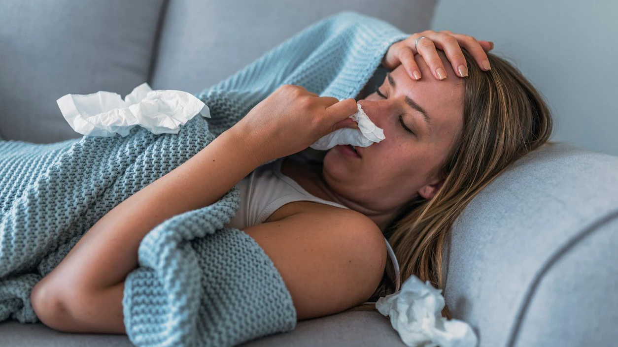 Sick Woman having Flu with Caught Cold. Sneezing into Tissue. Headache. Virus. Sick woman lying in bed with high fever. She is blowing nose. Photo of Young female Lying On Bed covered with a blanket  Infected With Allergy holding her head.