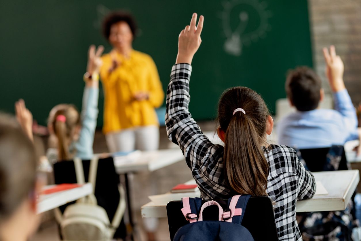 Back view of elementary student raising arm in order to answer a questing during a class.