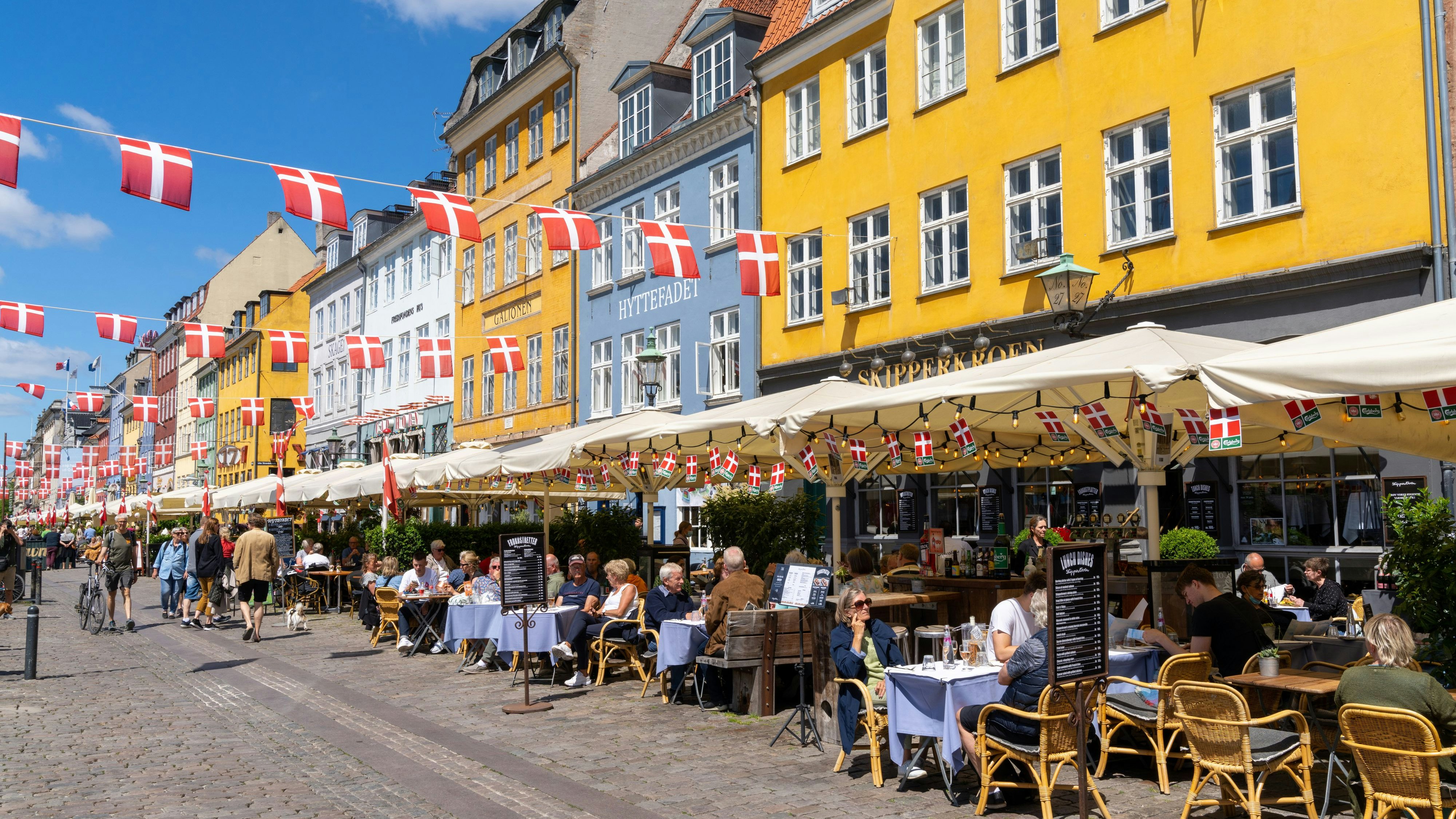 Copenhagen, Denmark - 13 June, 2021: people enjoy a summer day in the busy Nyhavn quarter on the waterfront in Copenhagen with many restaurants and bars and Danish flags above