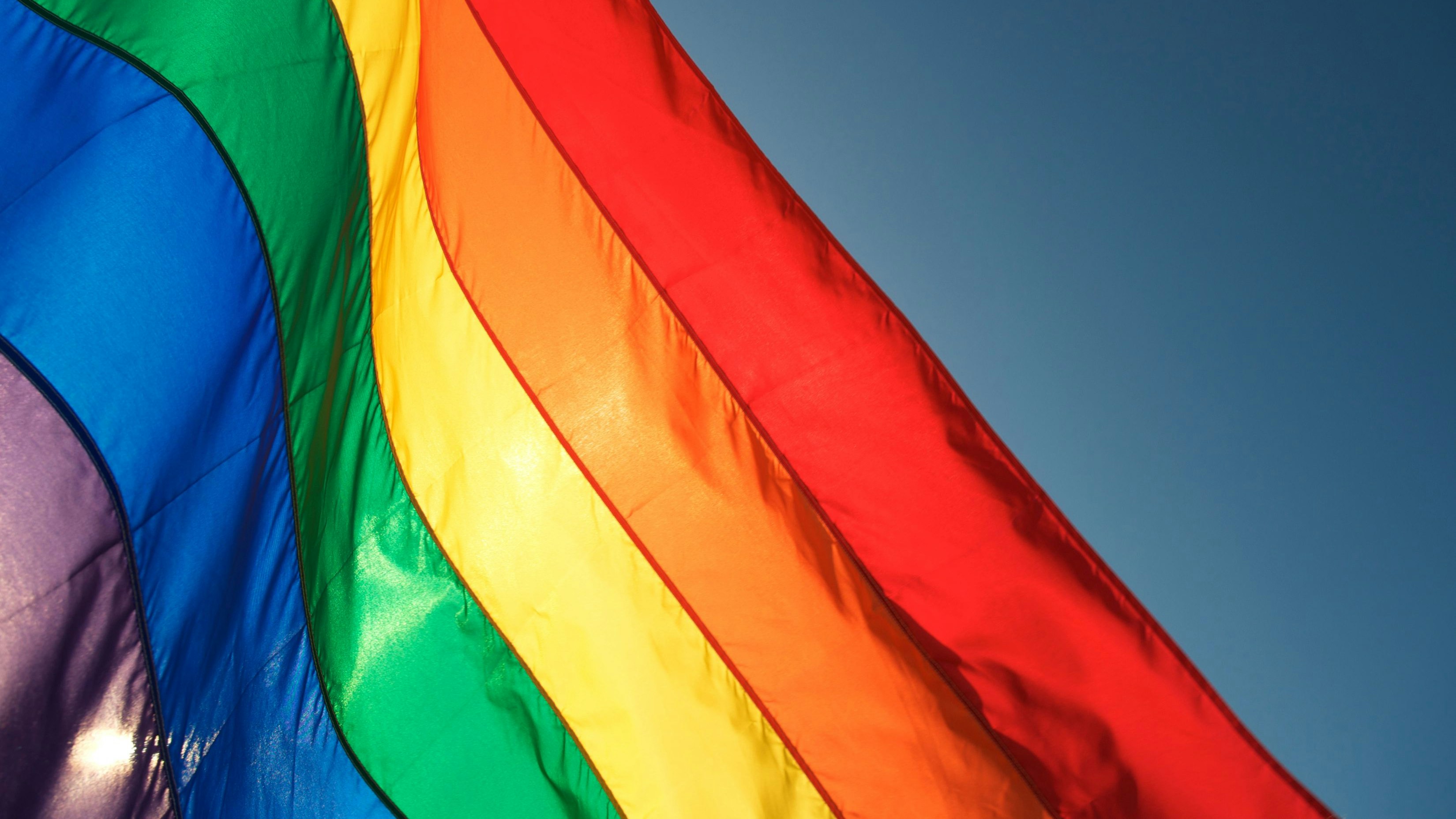 Rainbow flag glows in bright sun waving in blue sky. Some motion blur on top edge of waving flag.