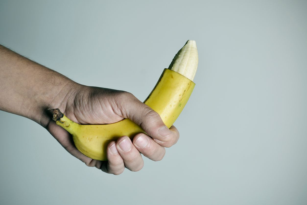 closeup of the hand of a young man with a banana with the tip of its skin removed, depicting a circumcised male member