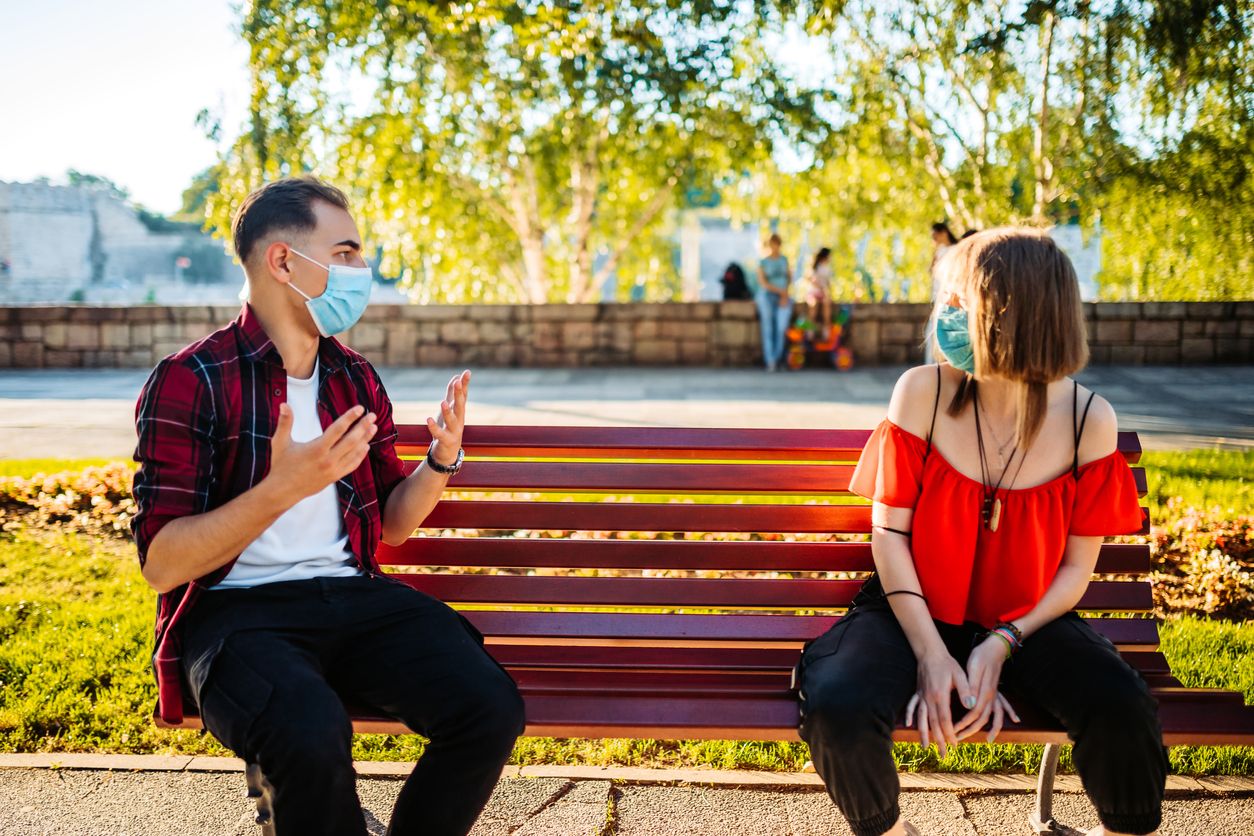 Cute Caucasian teenage couple on a date with protective face masks. Sitting on park bench and talking.