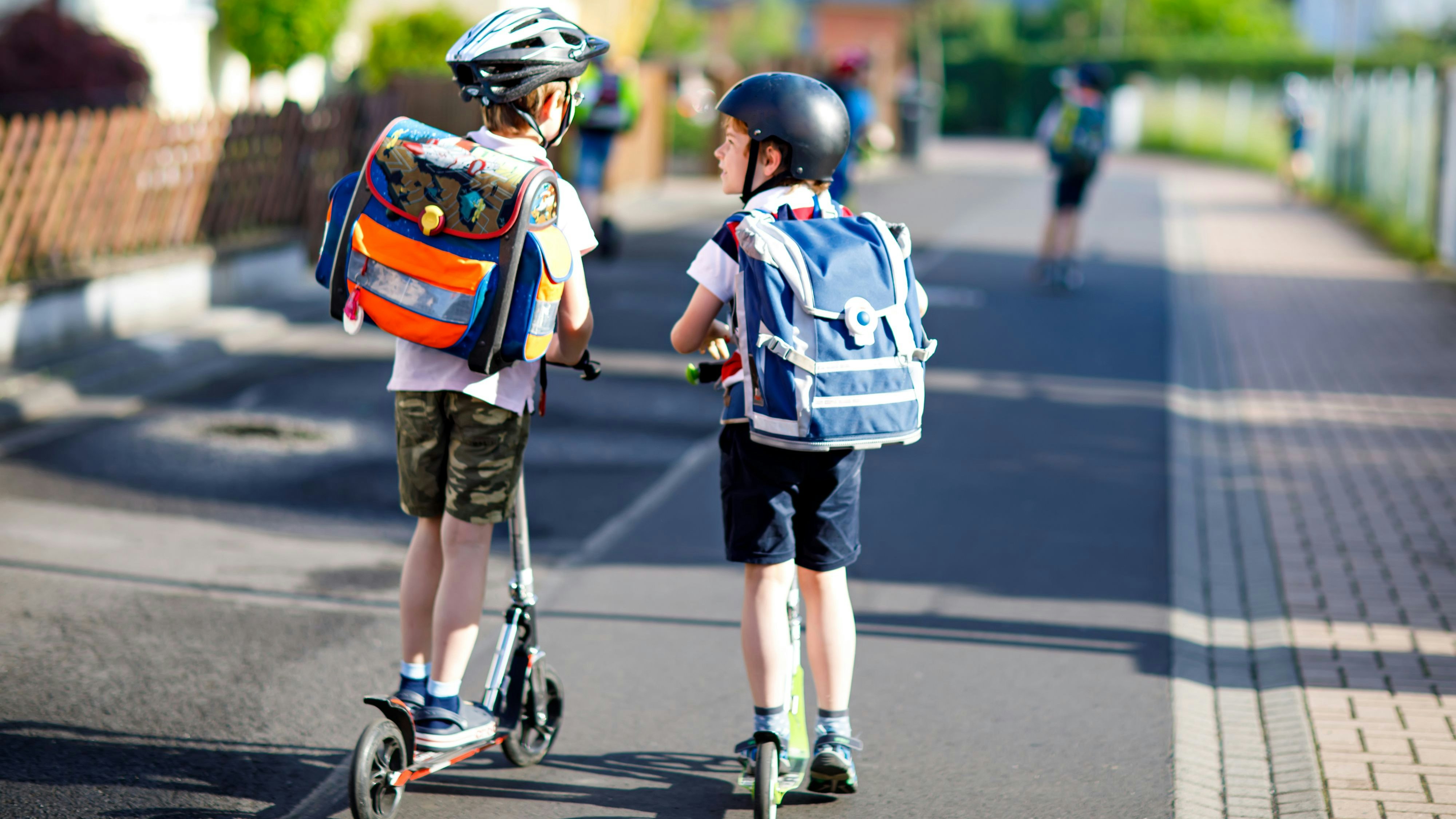 Two school kid boys in safety helmet riding with scooter in the city with backpack on sunny day. Happy children in colorful clothes biking on way to school. Safe way for kids outdoors to school.