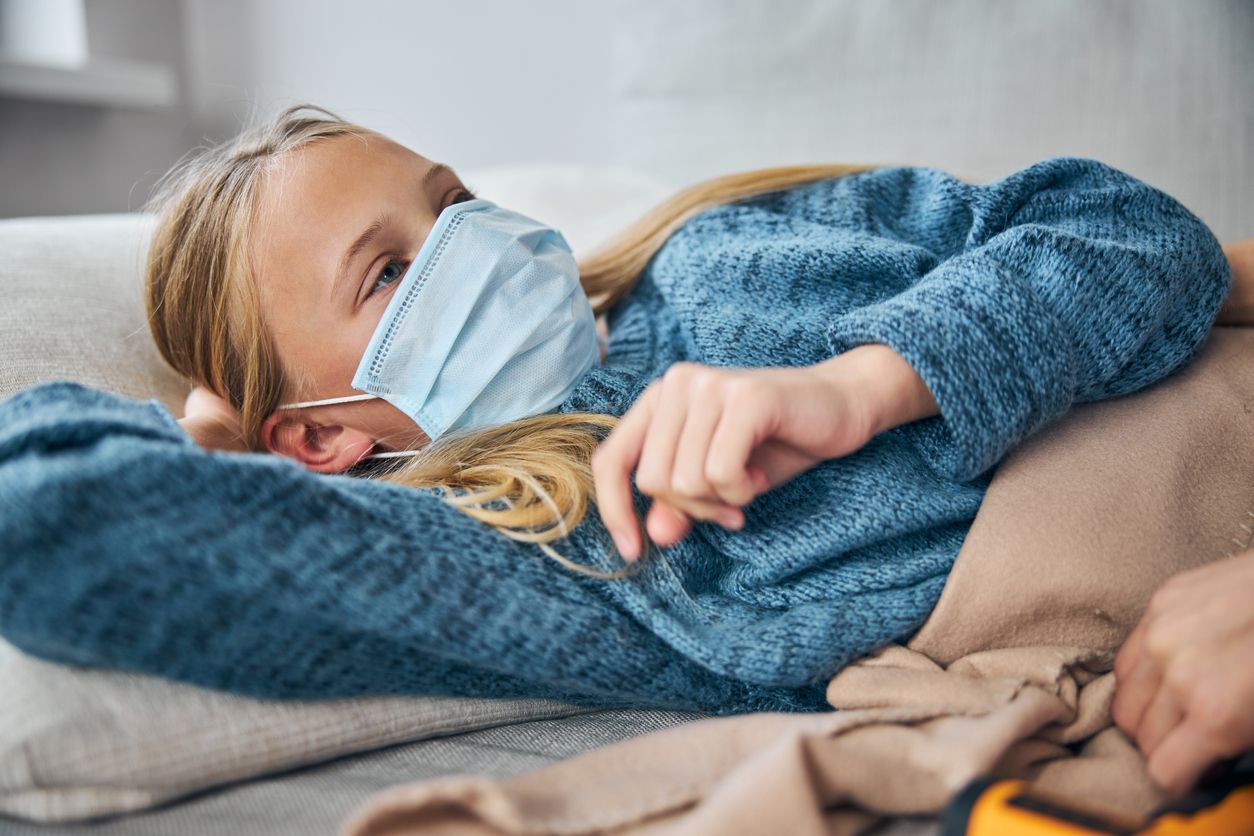 Low-spirited female child in a face mask lying on the sofa at home in broad daylight