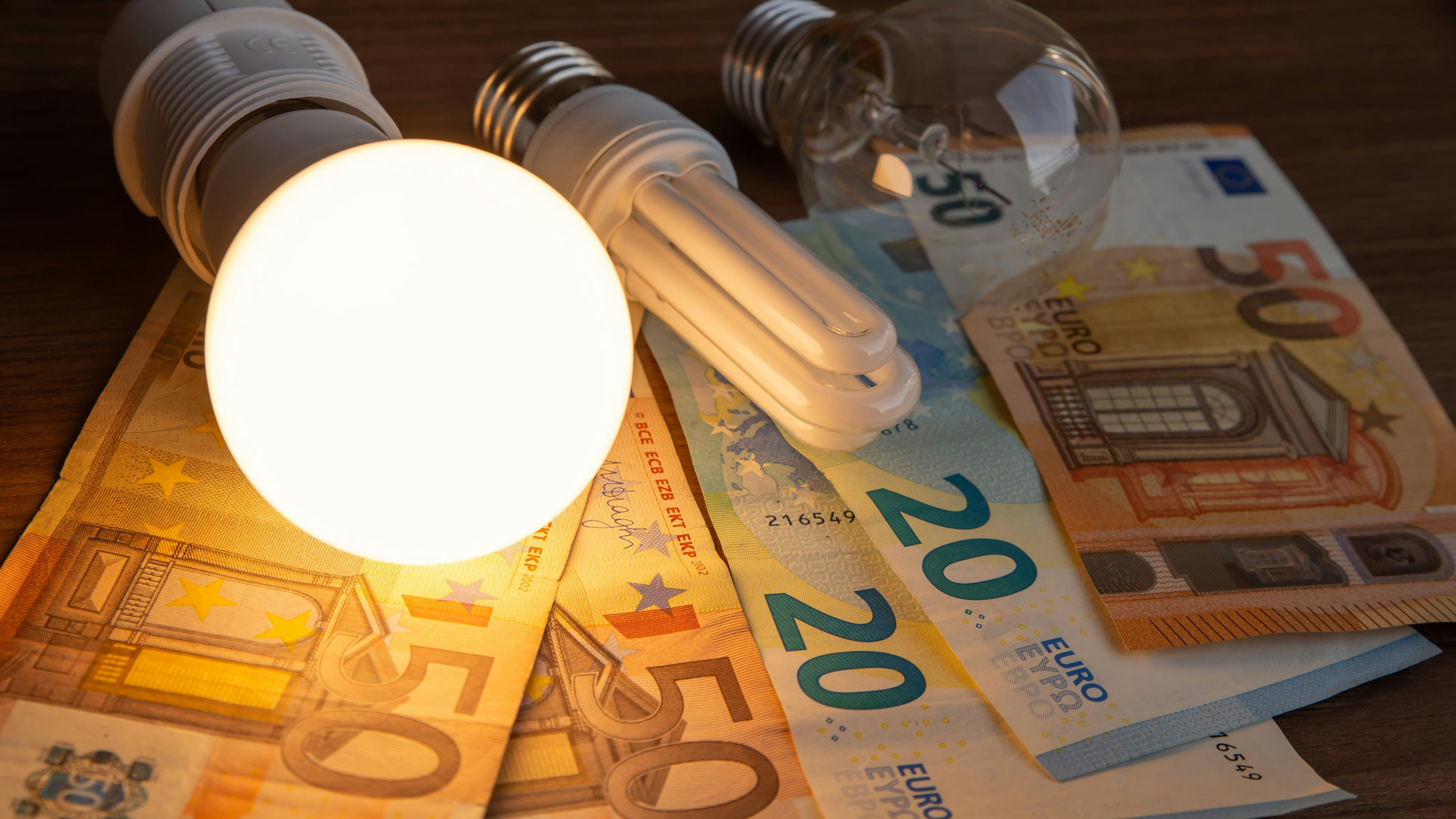 LED bulb on, with fluorescent bulb and incandescent bulb, with bills and coins next to it. Energy costs and evolution of light bulbs.