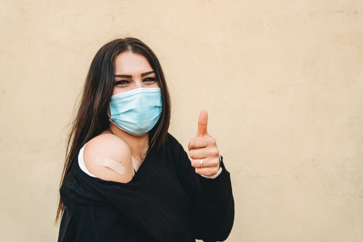 Young woman proudly shows her arm where she just got a vaccine. She's against a brown background wall.