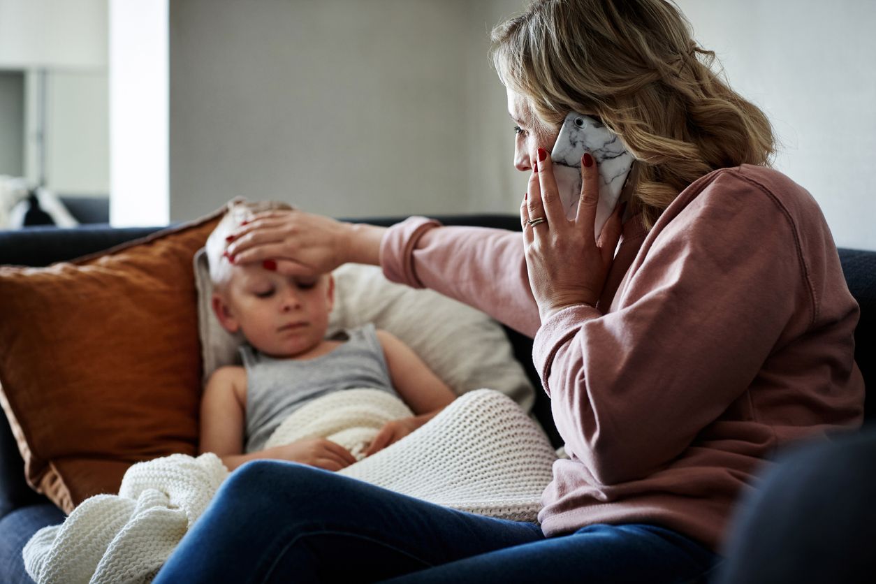 Shot of a mother wearing a surgical mask making a phone call while aiding to her sick young son at home