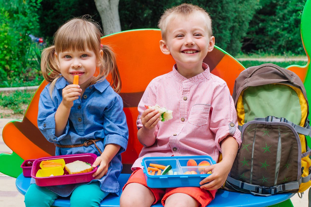 Little boy and girl preschoolers eating from their lunch boxes sitting outdoor