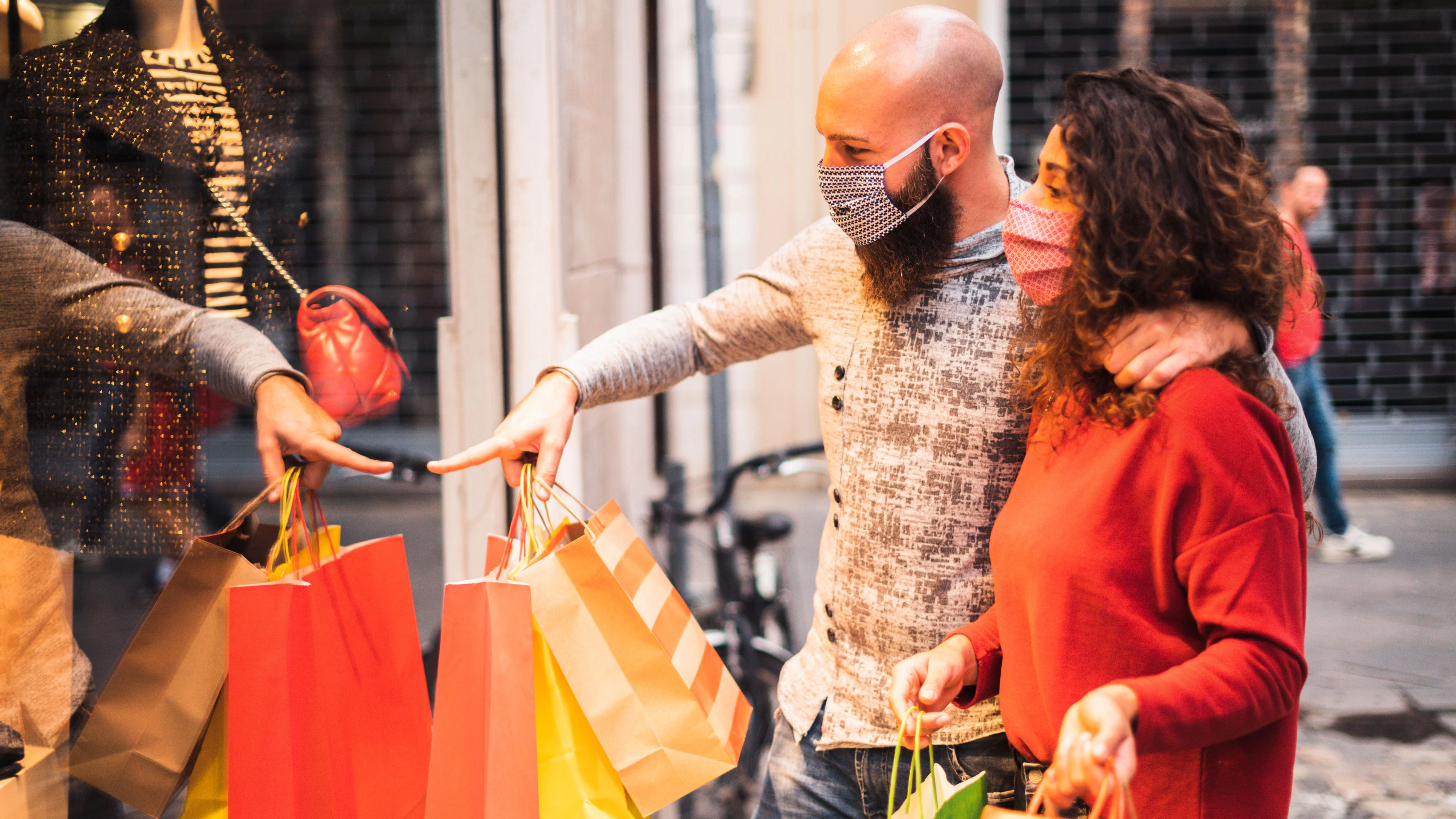Pretty young man pointing to shop window to show clothing item his likes to his girlfriend - Beautiful young couple enjoying in shopping, having fun together, with the face mask - Consumerism, love, dating, new normal, lifestyle concept
