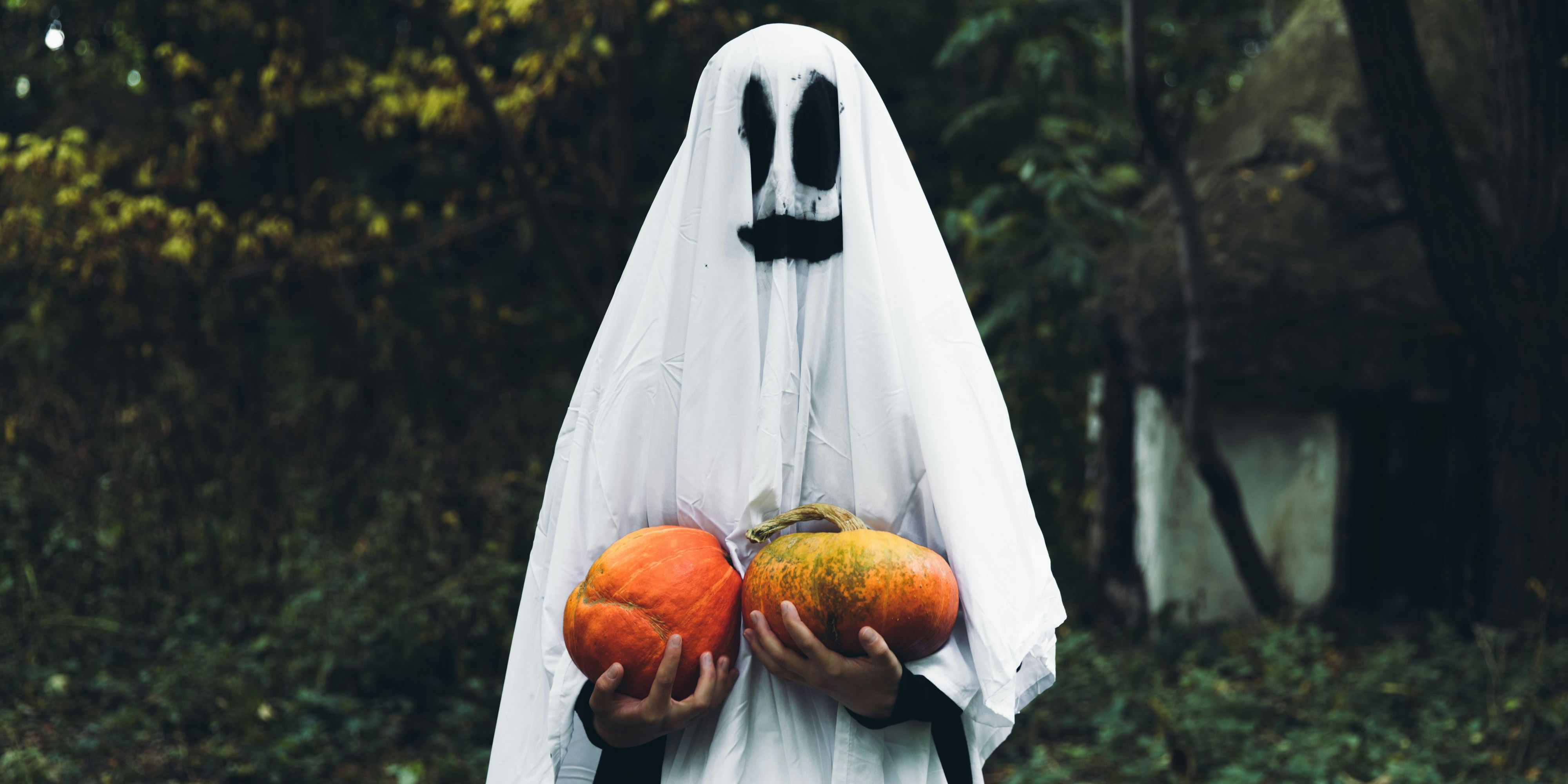 Woman dressed like a spooky smiling ghost holding two beautiful orange pumpkins and staying in the scary forest near abandoned house celebrating Halloween