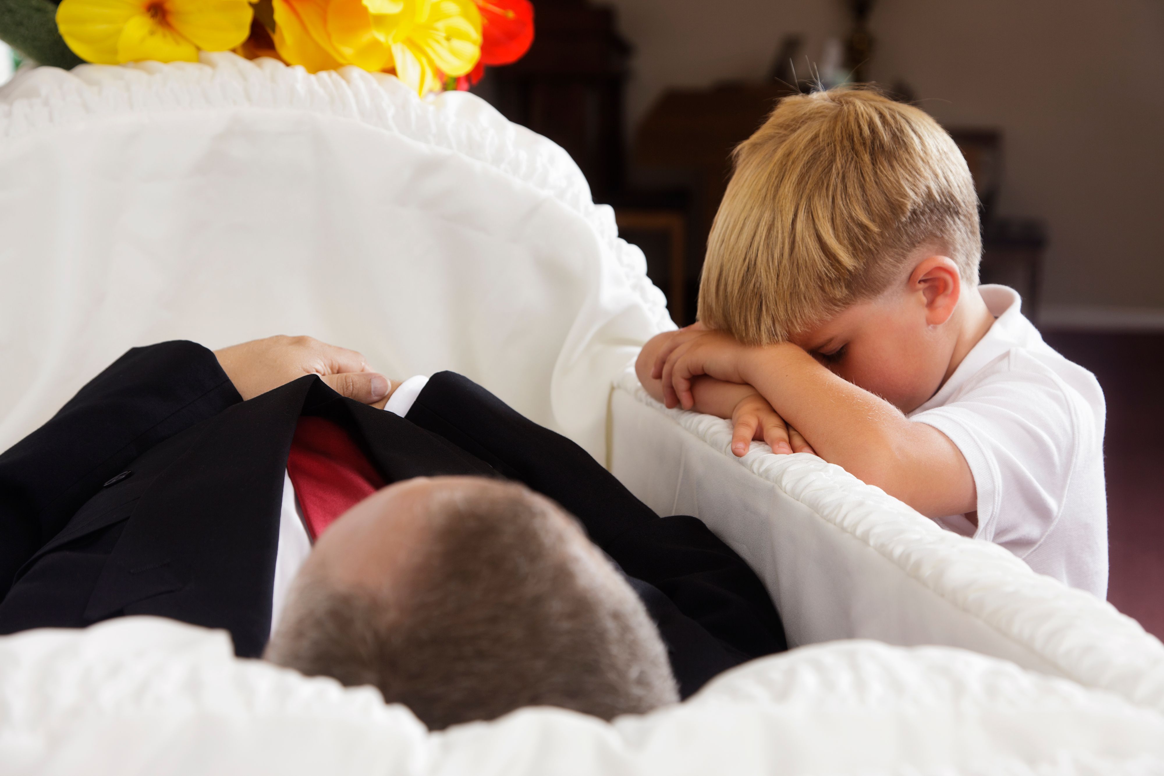 A sad little boy kneeling beside an open casket at a funeral.