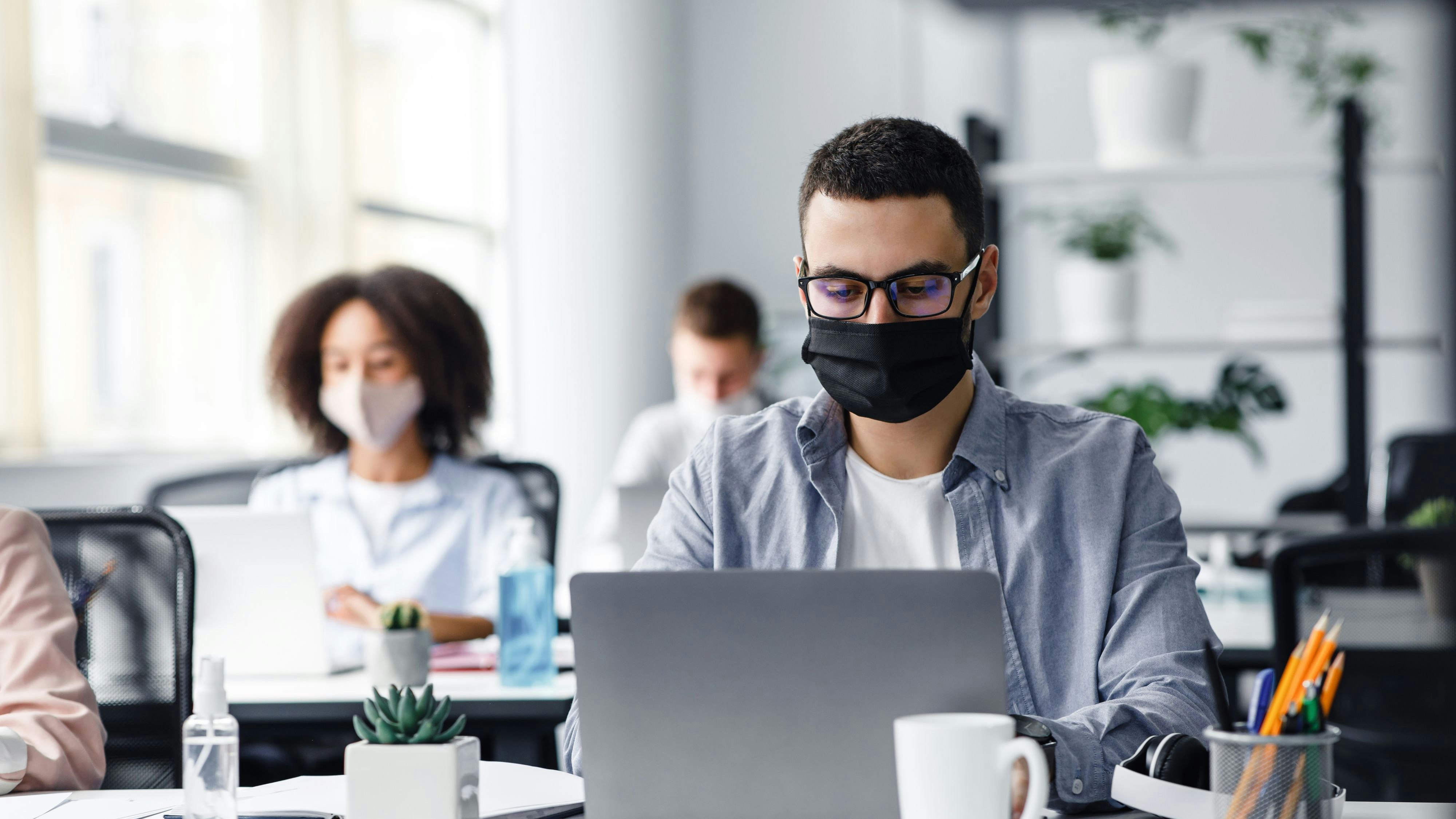 Office center workers are protected from virus outbreak during covid-19 epidemic. Young hipster man in glasses and protective mask works at laptop, with colleagues at workplaces in interior, free space