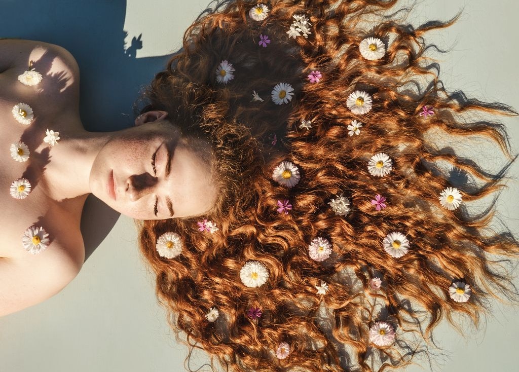 Close up portrait of young ginger curly girl with flowers in hair above. Top view. Concept of spring and summer youth happiness.