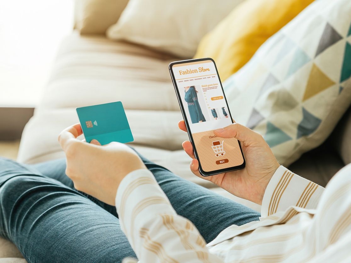 Stock photo of a woman buying a dress online with the phone and a credit card from the sofa at home. E-commerce concept