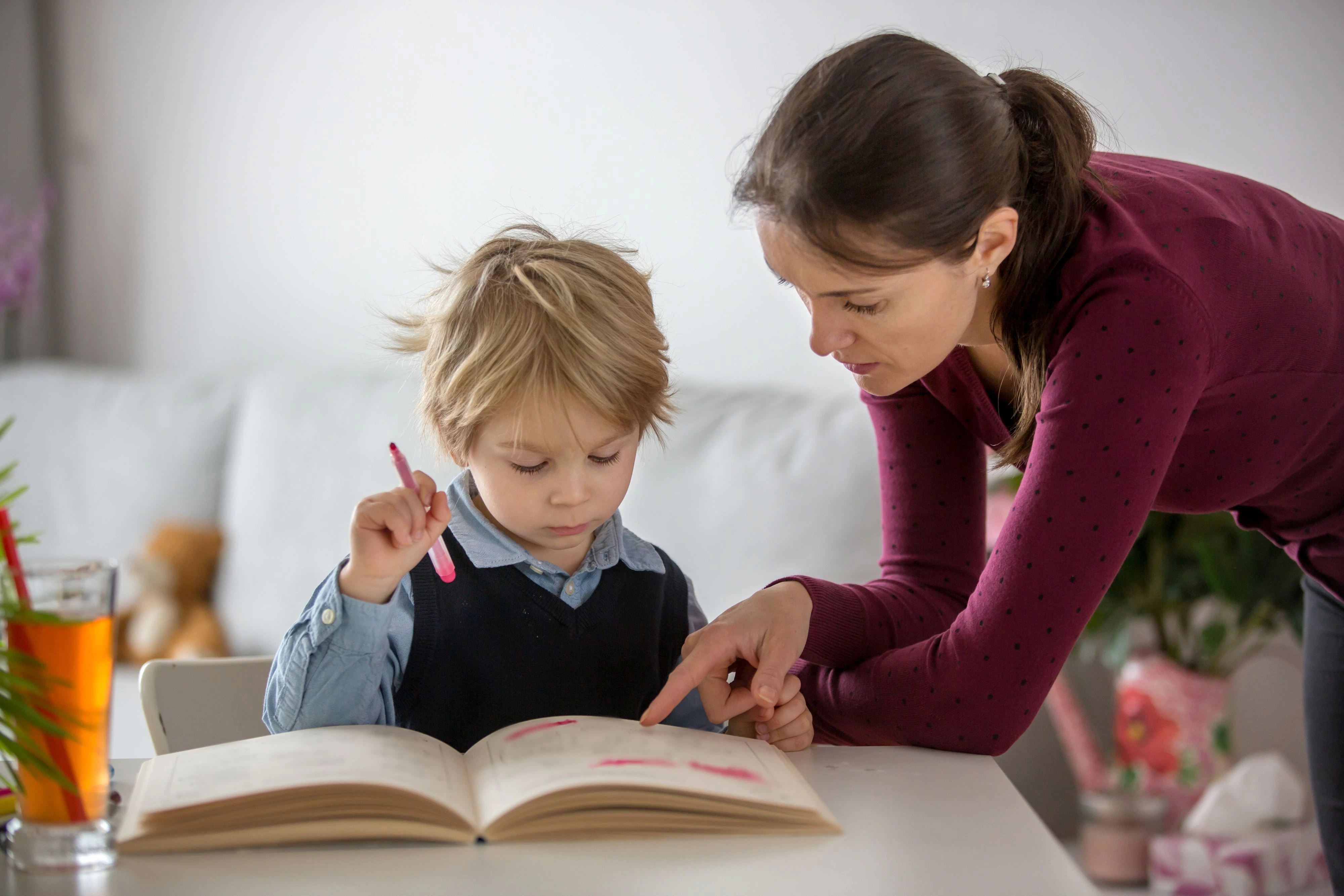 Cute preschool child, blond boy, filling some homework in a work book and coloring, mother helping him at home