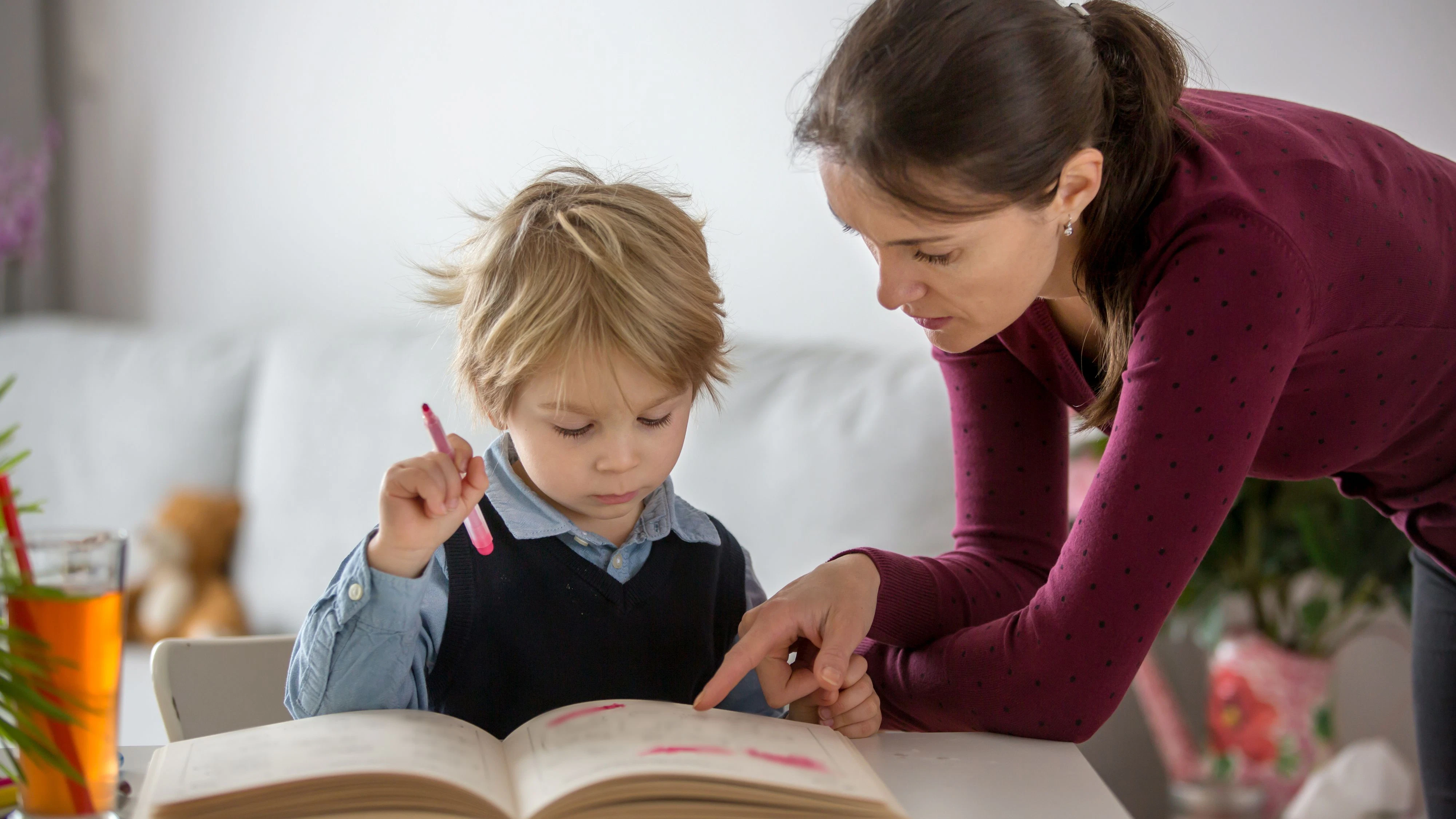 Cute preschool child, blond boy, filling some homework in a work book and coloring, mother helping him at home