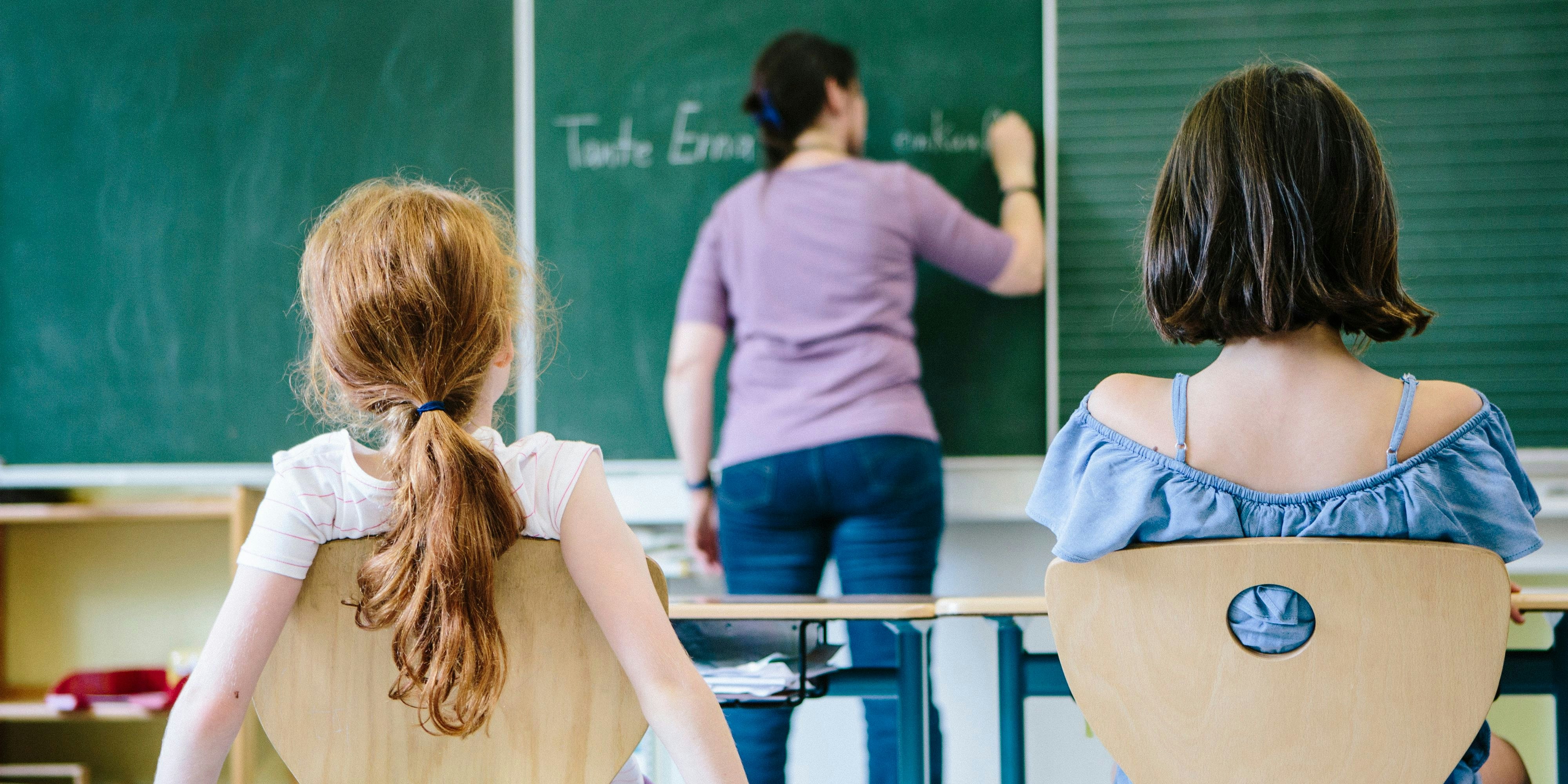 a female teacher writes at the blackboard. rear view of two girls in front.