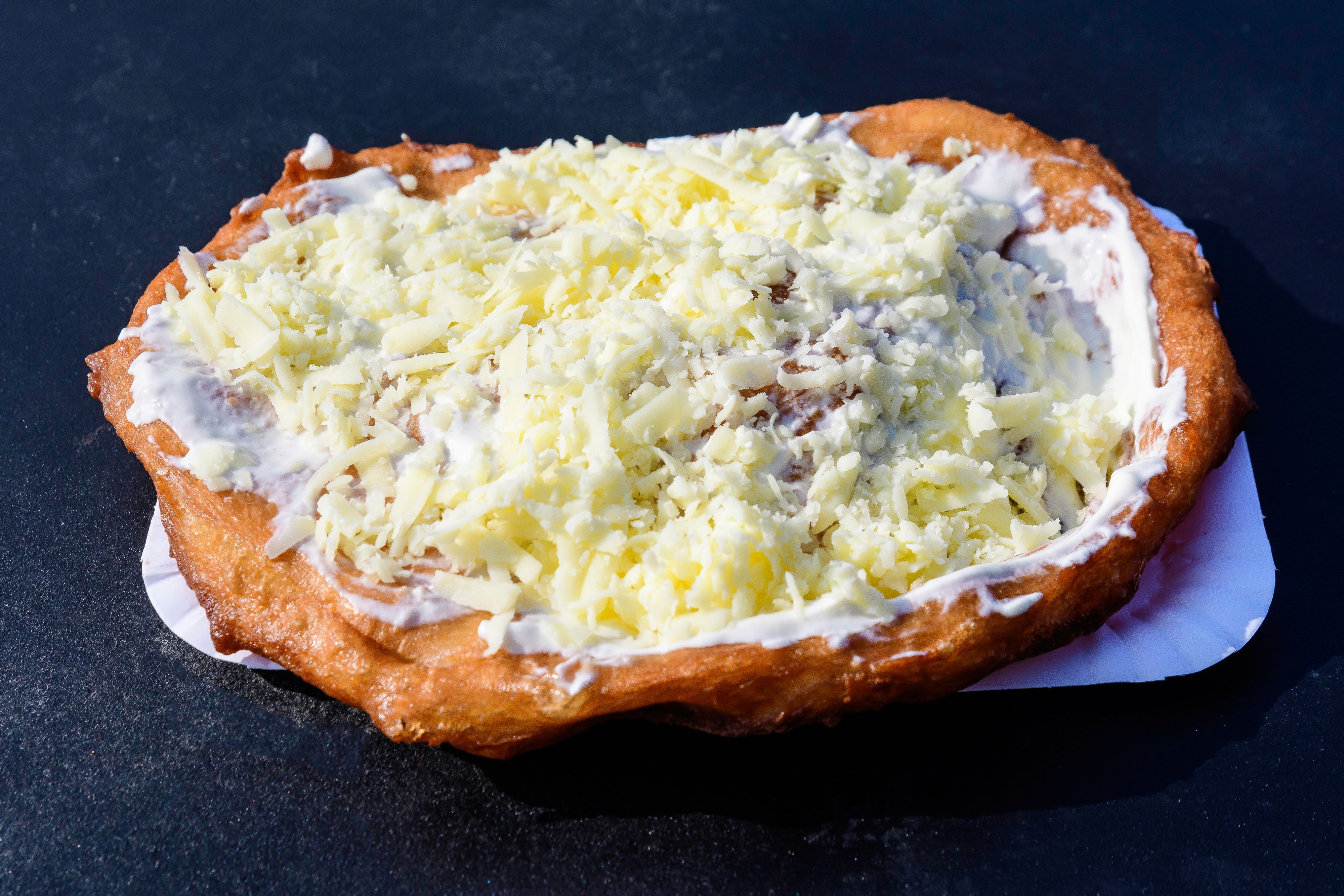 Close up of a langos, typical Hungarian food specialty, with cream and cheese on a white paper on a  black metal table, ready for take away, deep fried donut in direct sunlight at a food market