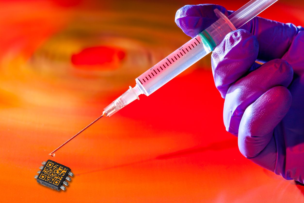 Vaccine injection syringe held by healthcare worker's hand in blue surgical protective glove, above microchip tracker in conspiracy theory concept, concerning COVID-19 and COVID-20 illness. Concept also applies to other vaccines and diseases. Copy space provided.  Note: The QR code and text on the vaccine bottle was generated by me and contains generic text: "SARS-CoV-2 Vaccine". The QR code on the microchip is also from me with text: "COVID-19 Vac Nr 1234567890".